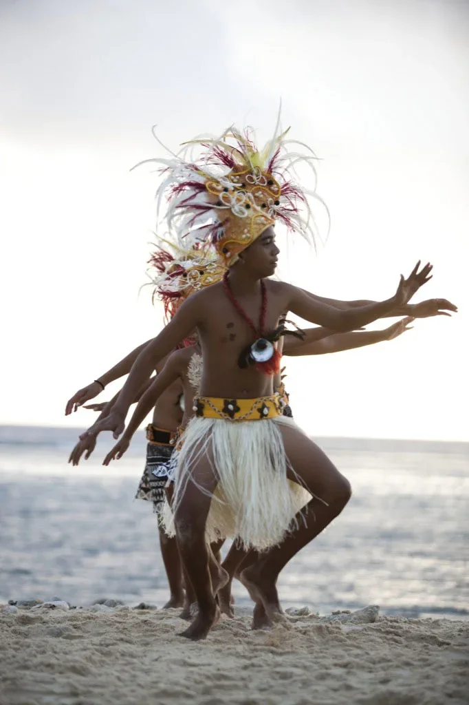 Traditional Polynesian dancers in grass skirts and feathered headpieces performing on the beach near the water at sunset.