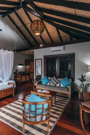 Interior of a tropical-style living room with a vaulted ceiling, wooden beams, a wicker pendant light, a sofa with blue and black pillows, a wooden armchair with a blue cushion, a striped rug, and dark wooden floors.