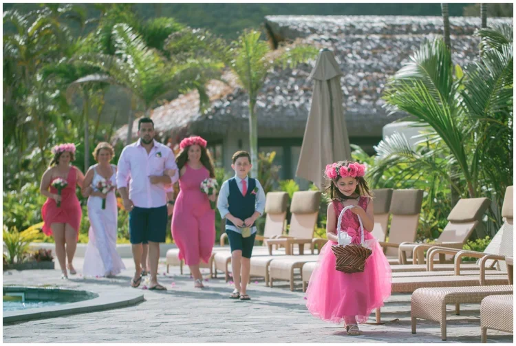 A young girl in a pink dress and floral crown holding a basket walking near a pool at a tropical resort, with a wedding party including bridesmaids and a bride in the background.