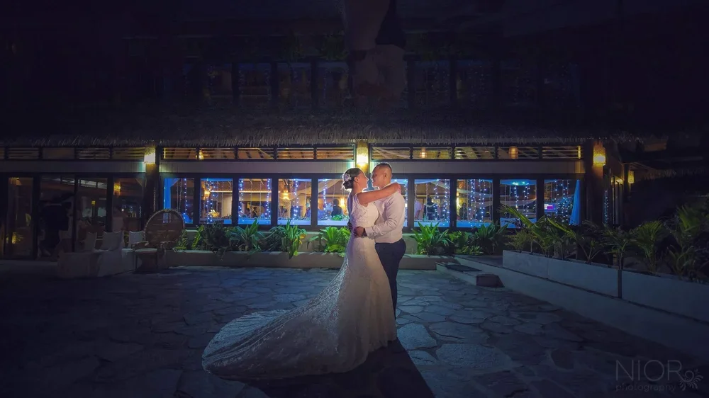 A bride and groom dancing outside at night, with a building decorated with lights and plants in the background.