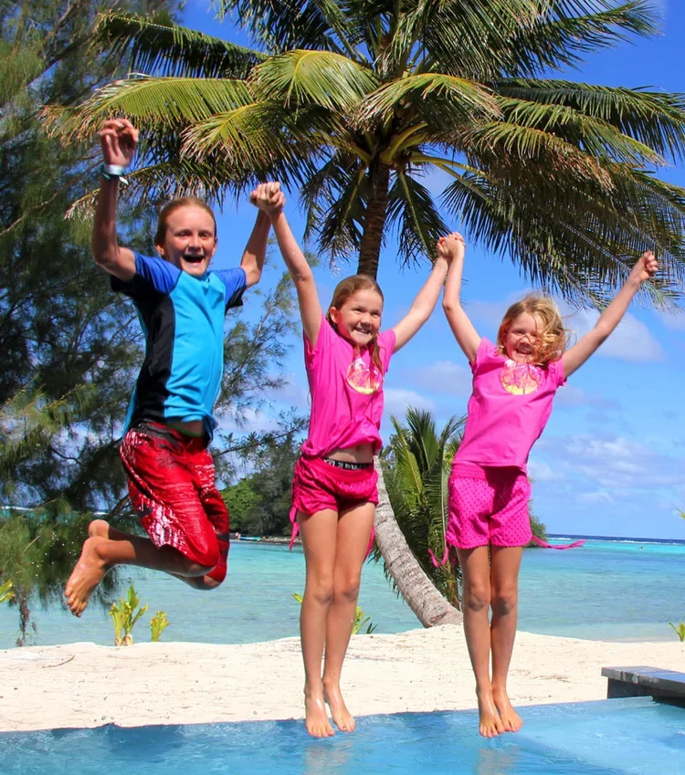 Three children jumping with arms raised on a tropical beach near a palm tree, bright blue sky, and ocean in the background.