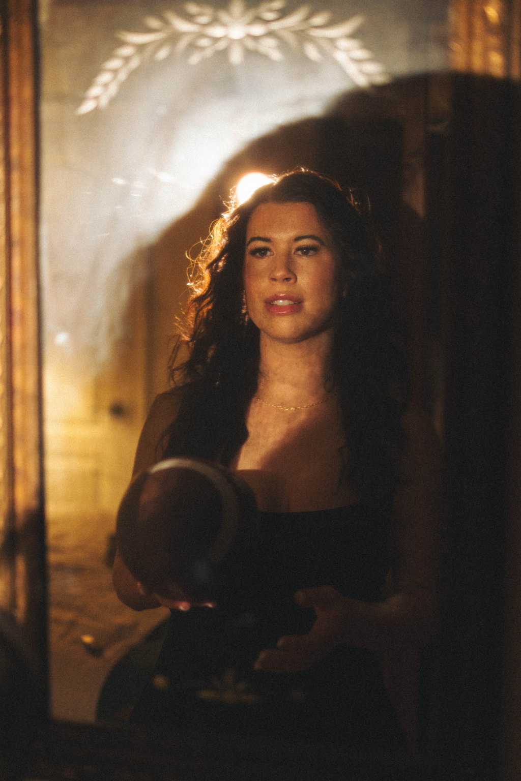 A woman with curly dark hair standing in front of a mirror, holding a hairbrush, with warm lighting illuminating her face.