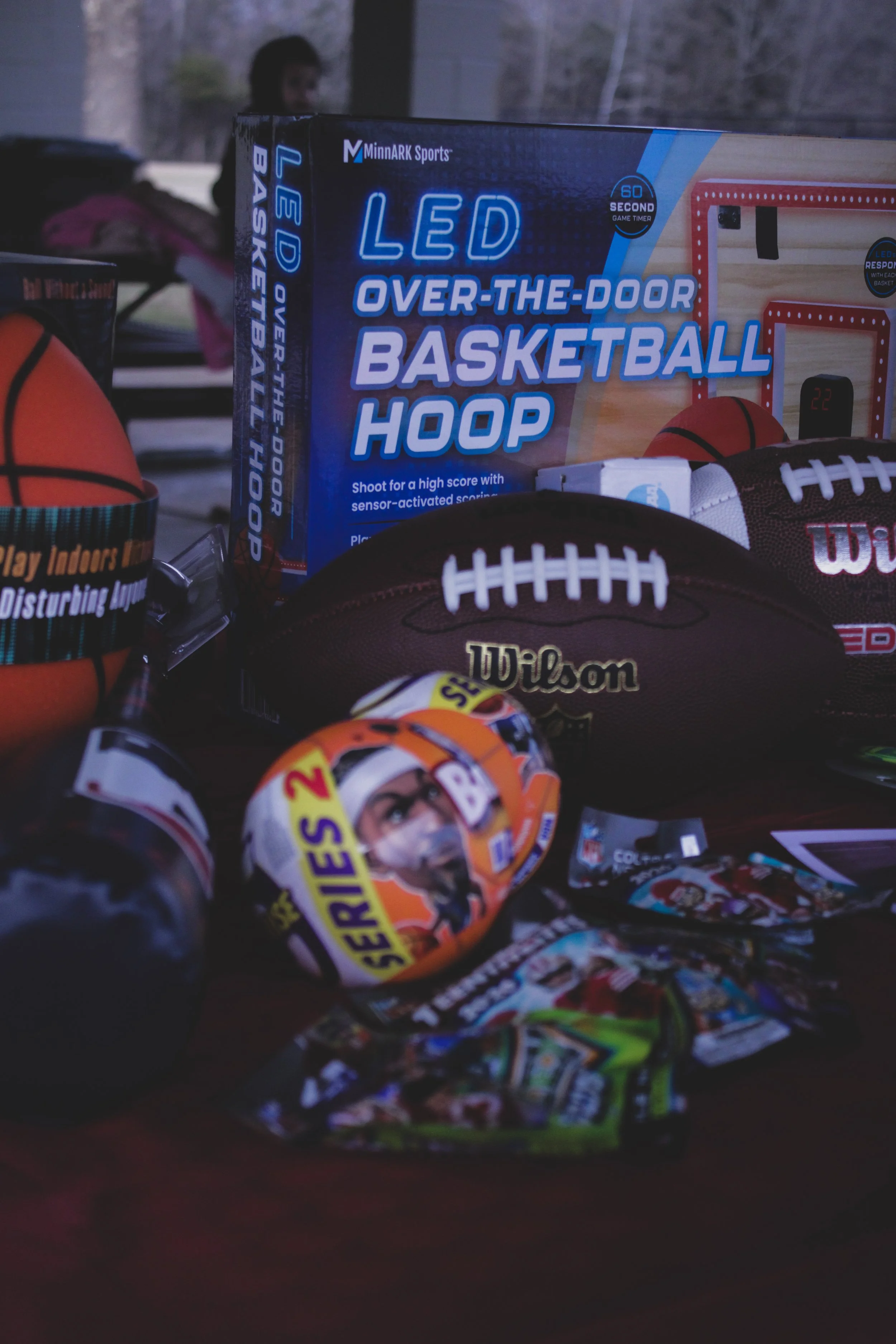 Box of LED over-the-door basketball hoop, several basketballs, toy cars, a Wilson football, and candy on a dark surface.
