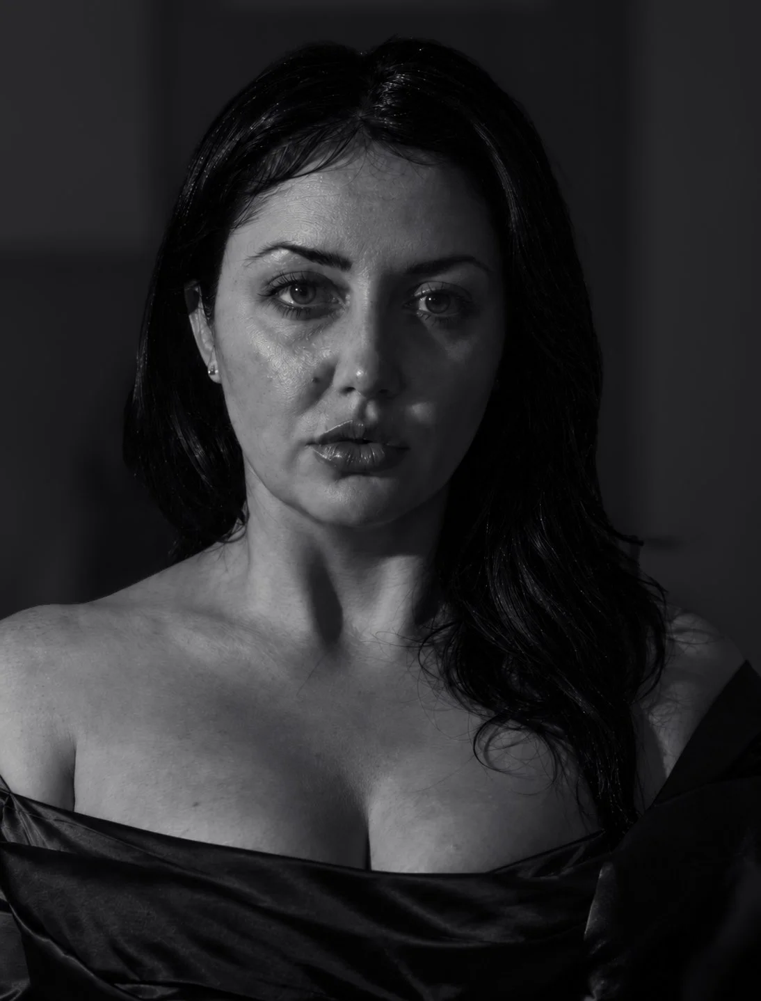 Black and white close-up of a woman's face and shoulders, with dark hair, makeup, and an off-the-shoulder garment.