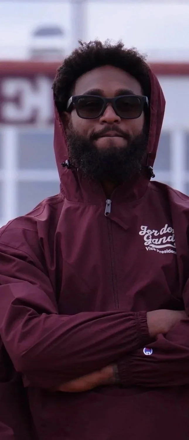 Man with curly hair and beard wearing sunglasses and a maroon Jordan Garden jacket standing with arms crossed.