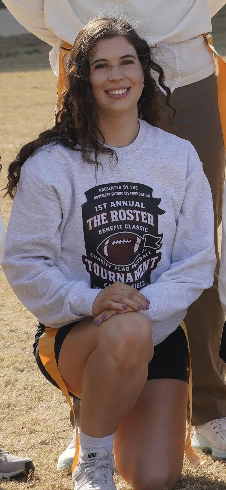 A young woman standing outdoors on a grassy field, wearing a sweatshirt, shorts, and running shoes, during daylight. She appears to be watching or waiting at an event, with other cars and trees in the background.
