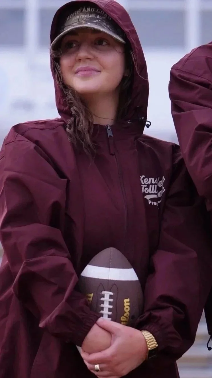 A woman wearing a maroon rain jacket and sporting a cap stands outdoors holding a football, with another person partially visible beside her.