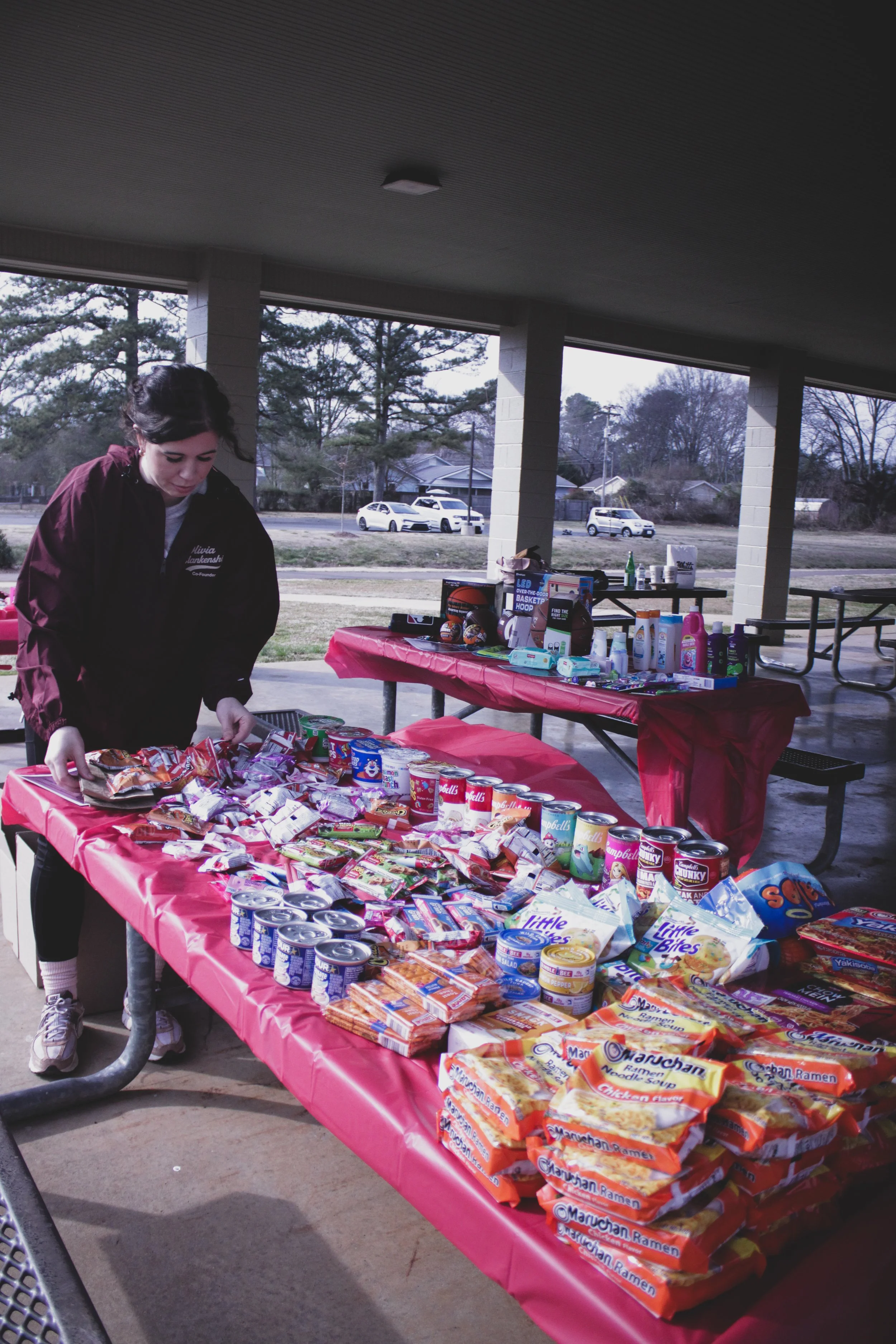 Person arranging snacks on a table with various food and beverage items during an outdoor event under a pavilion.