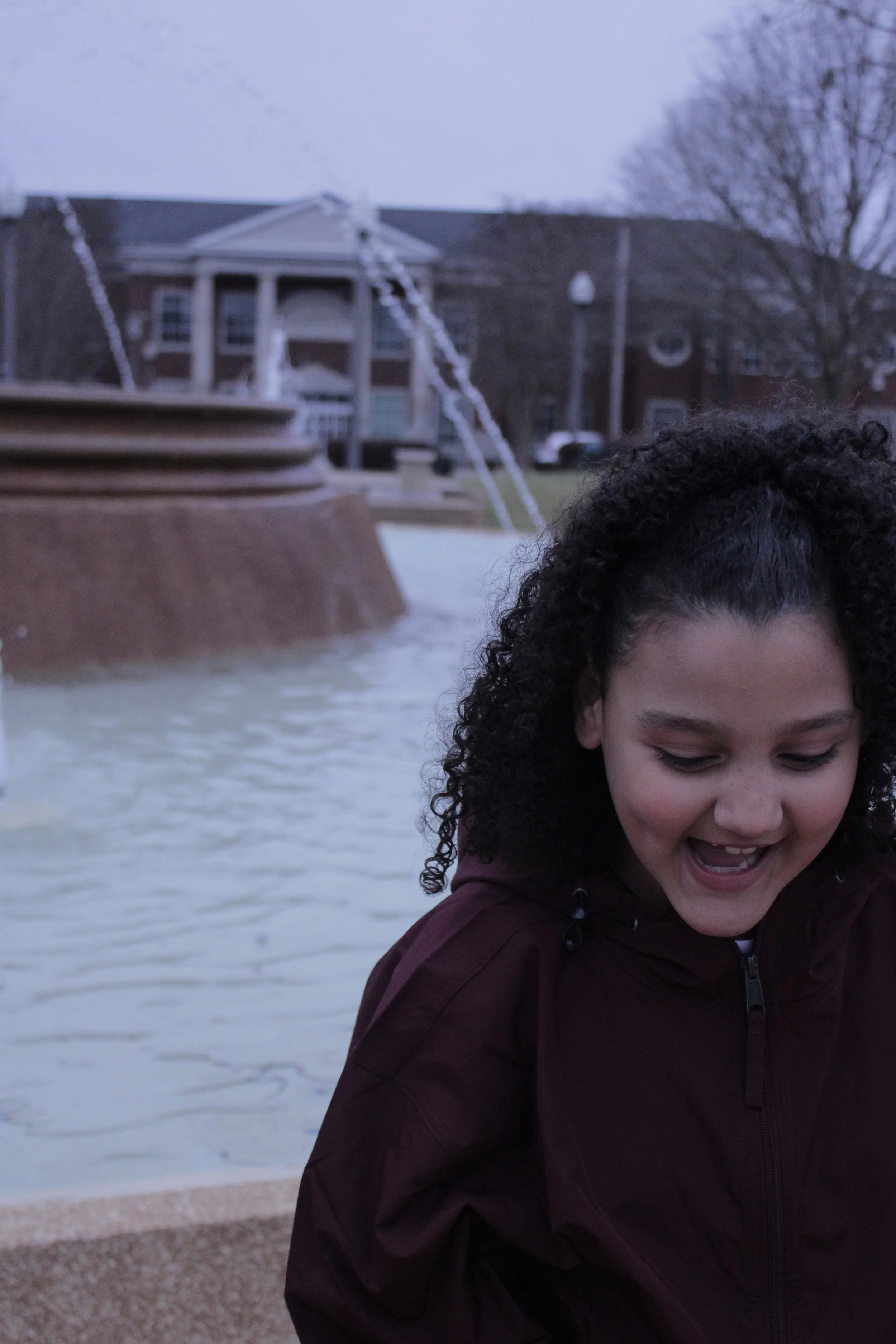 A young girl with curly hair smiling and looking down in front of a fountain with water spraying from it, in a park or public space with trees and buildings in the background.