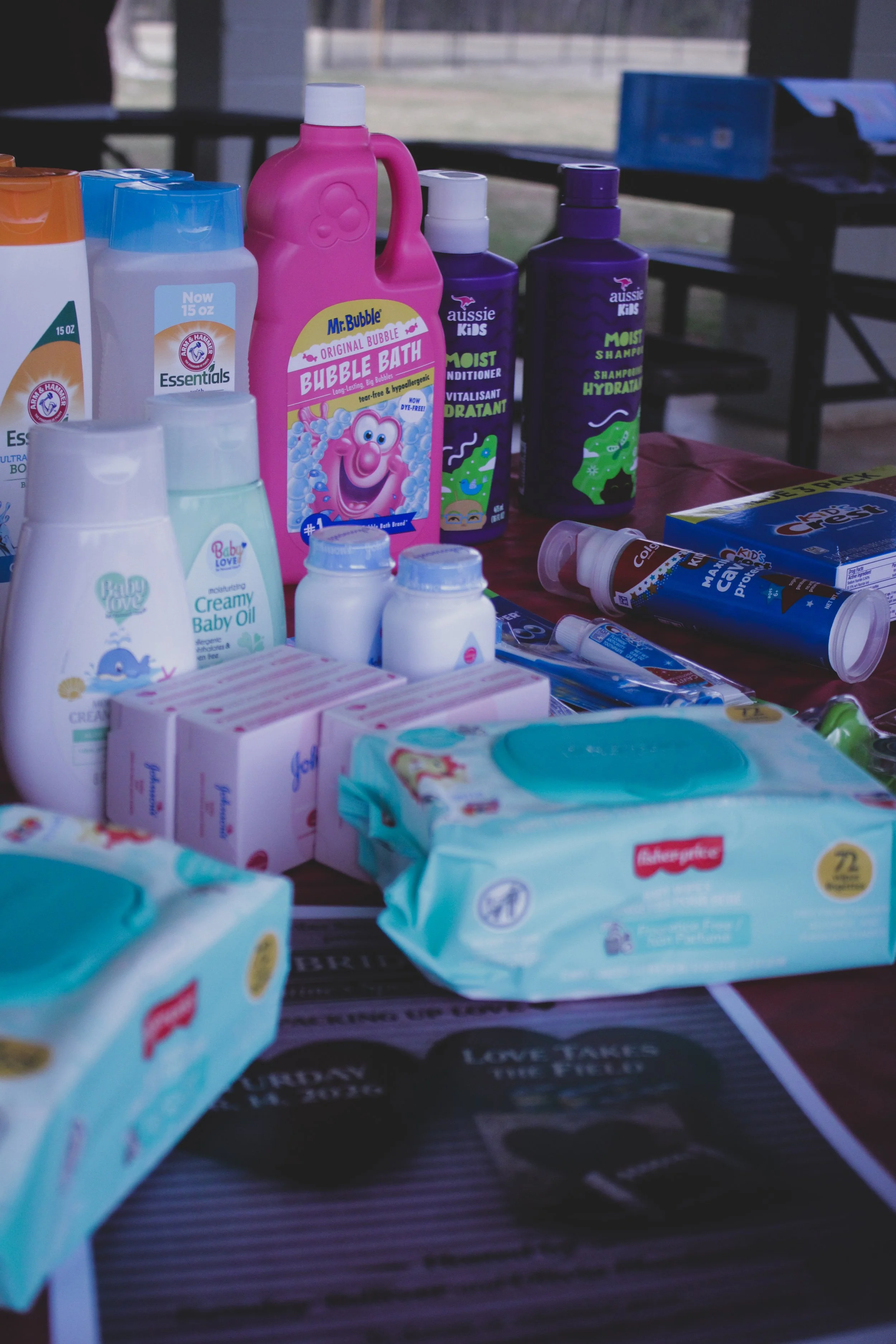A table filled with various baby and children's toiletries including bubble bath, shampoo, conditioner, baby oil, creams, wipes, and toothpaste, with a park visible through windows in the background.