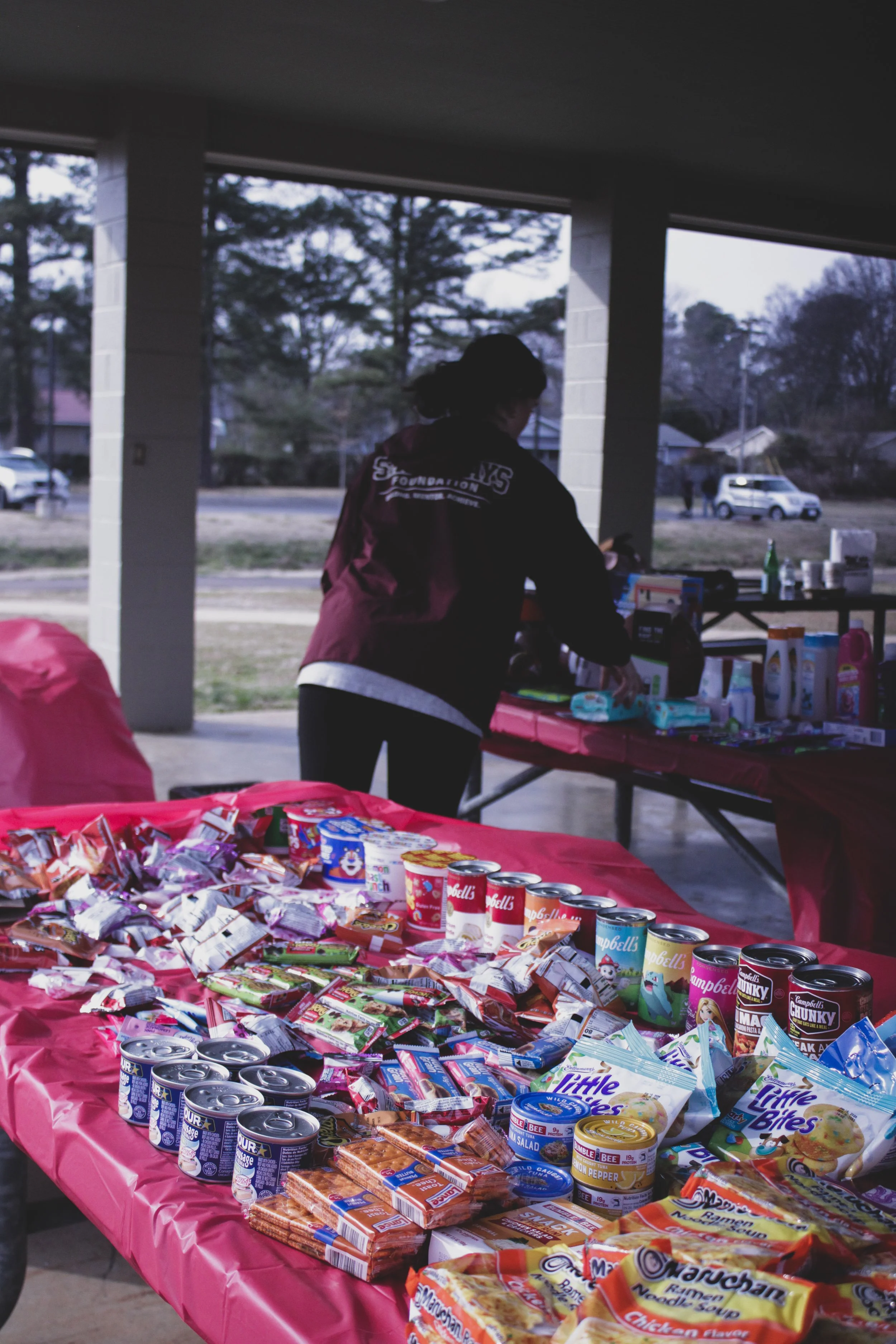 A table covered with snacks and canned drinks at an outdoor gathering, with a woman organizing items.