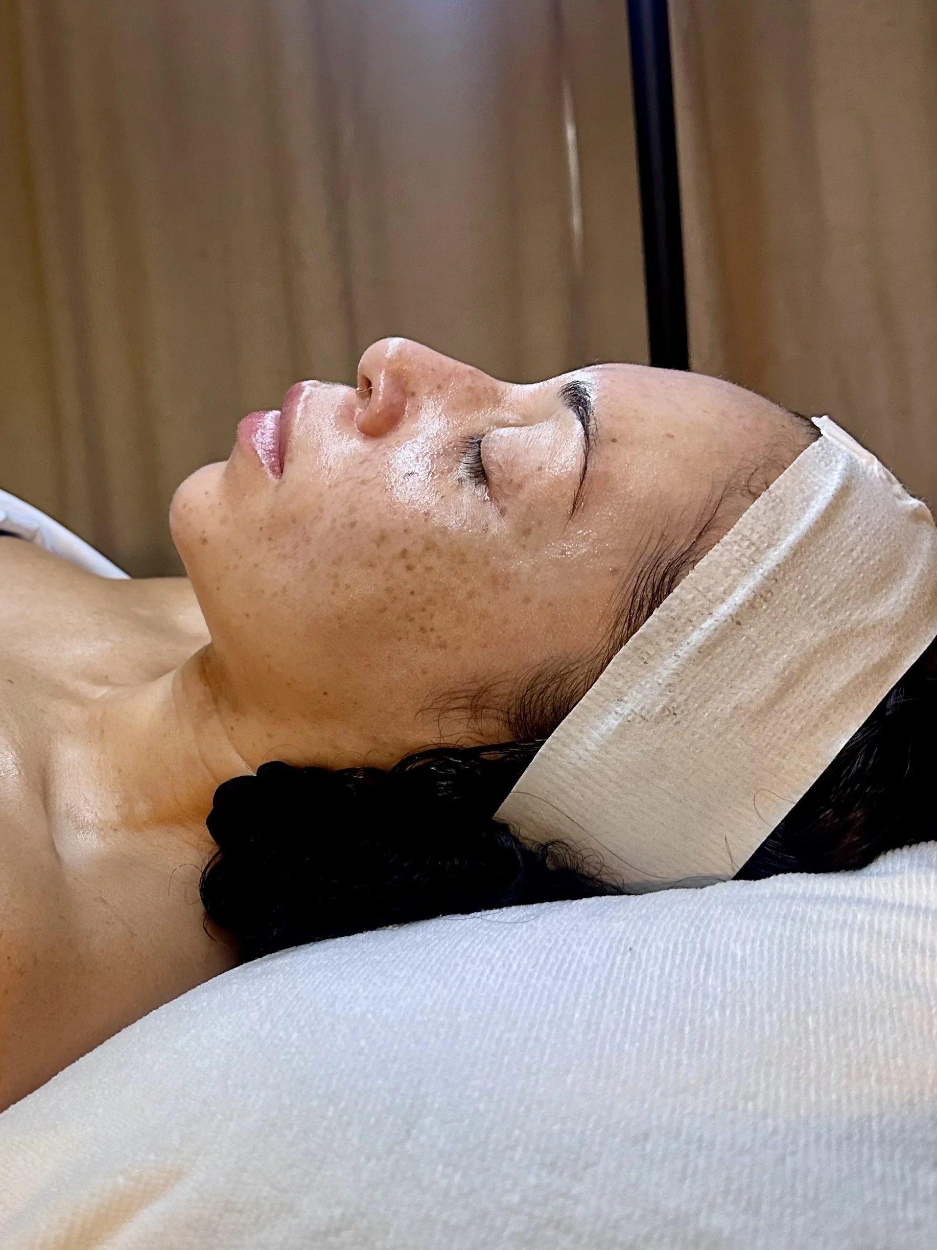 Woman lying down with eyes closed, wearing a headband, in a relaxing or spa setting with wooden background.
