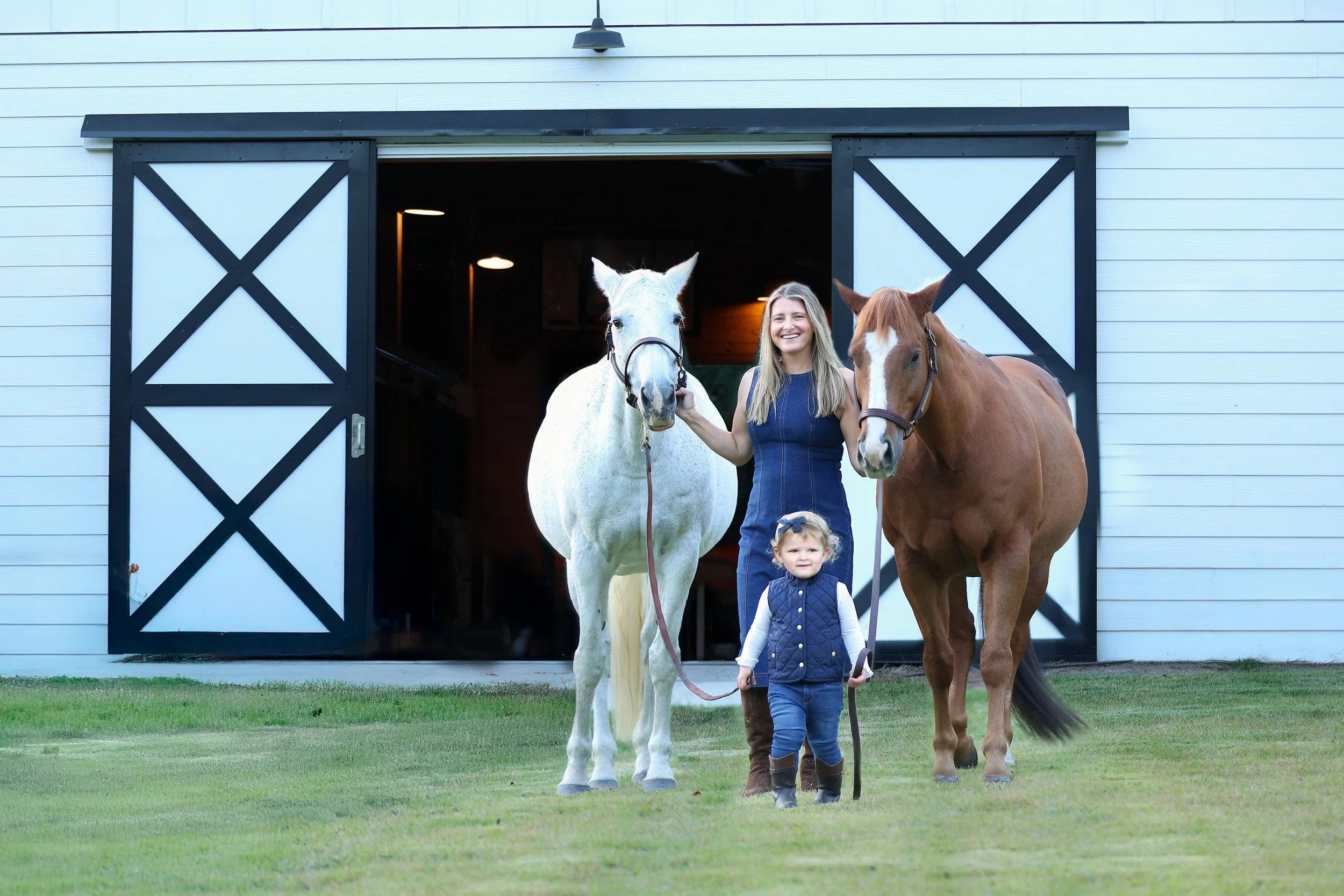 A woman and a young girl standing in front of a barn with two horses, holding their reins.
