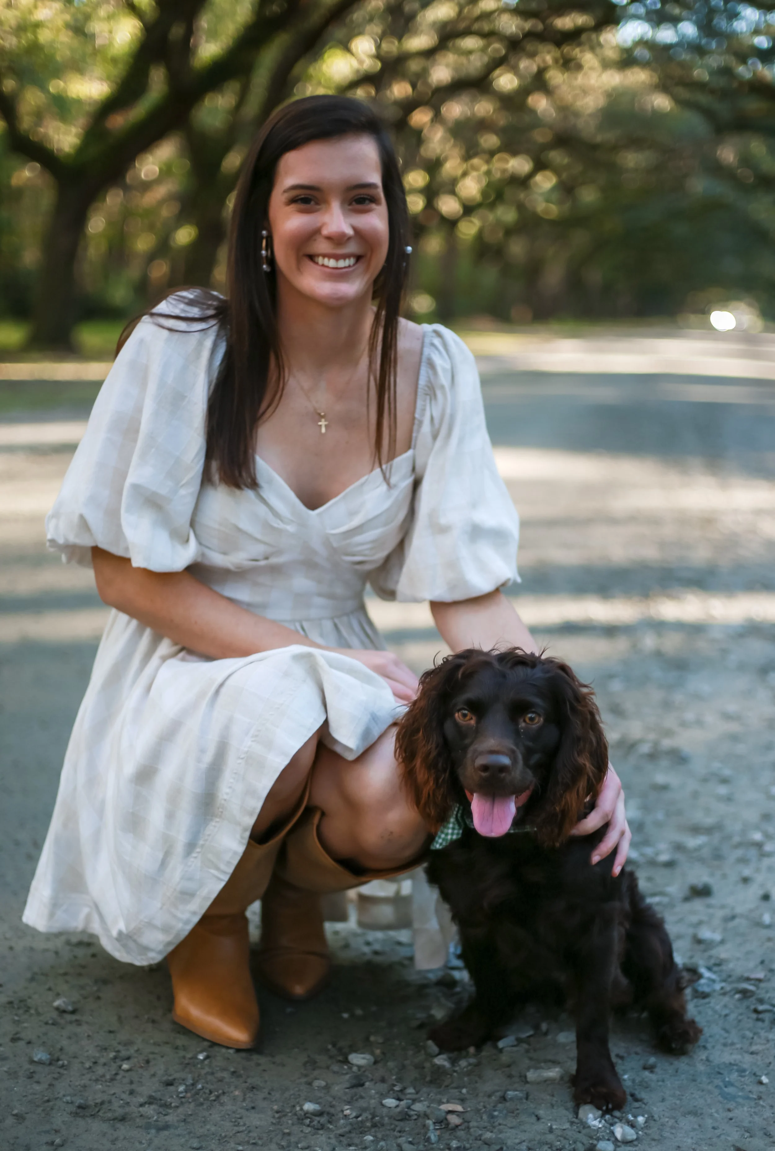 A young woman with long dark hair, wearing a white dress and cowboy boots, kneels next to a black Cocker Spaniel wearing a green bandana in a park with trees in the background.