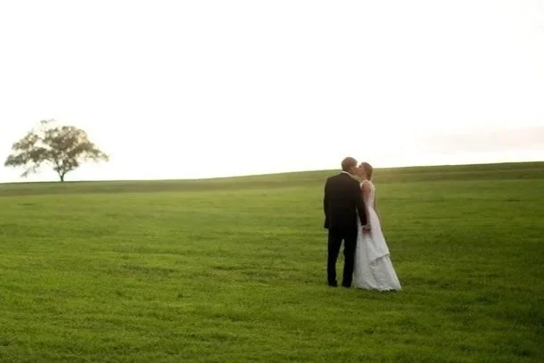 A newlywed couple in Western attire standing outdoors during sunset. The bride is in a white wedding dress and large white hat, holding a bouquet, while the groom is in a black suit with a black cowboy hat, placing a ring on her finger.