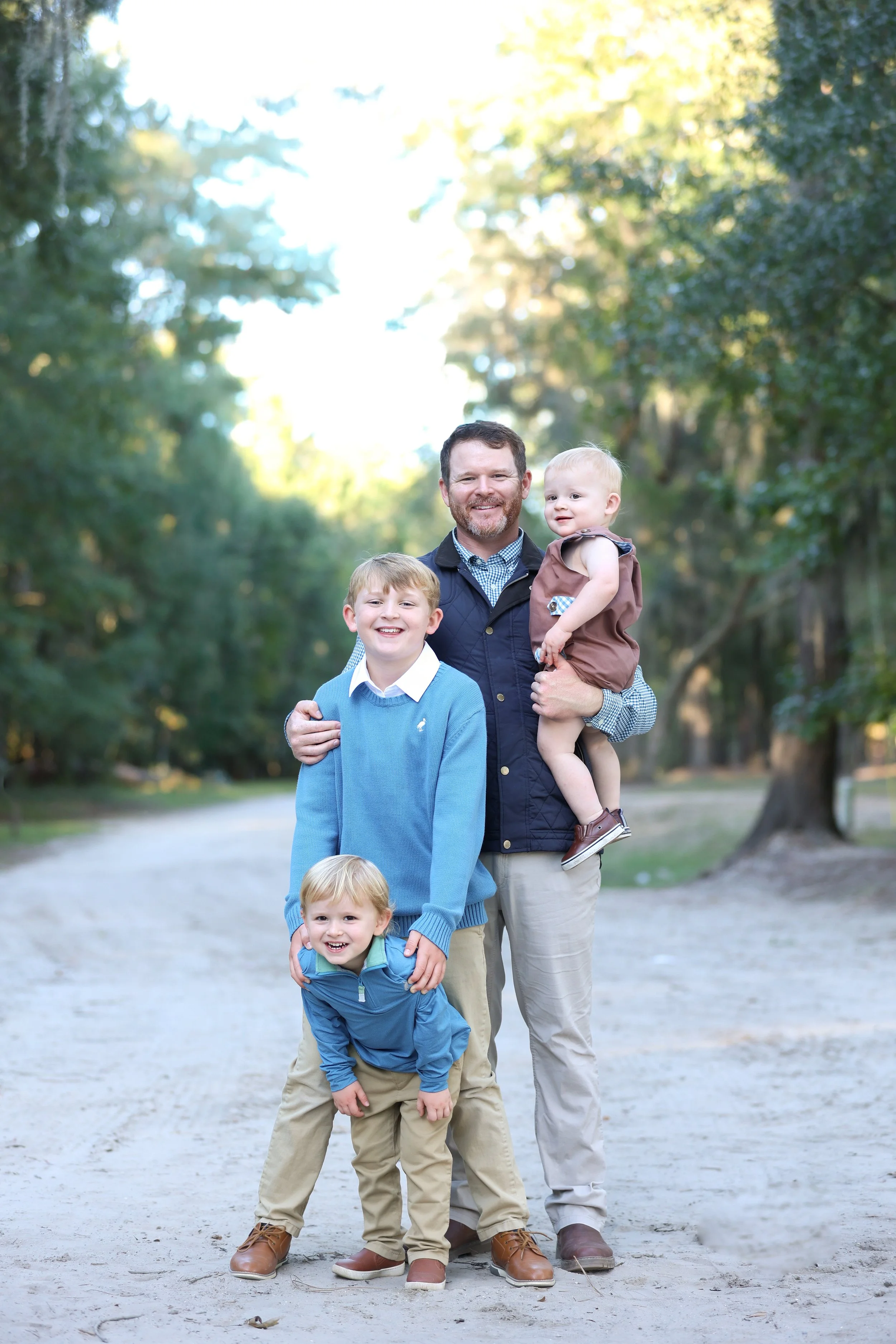 A man with three young children outdoors on a dirt path surrounded by trees, smiling at the camera.