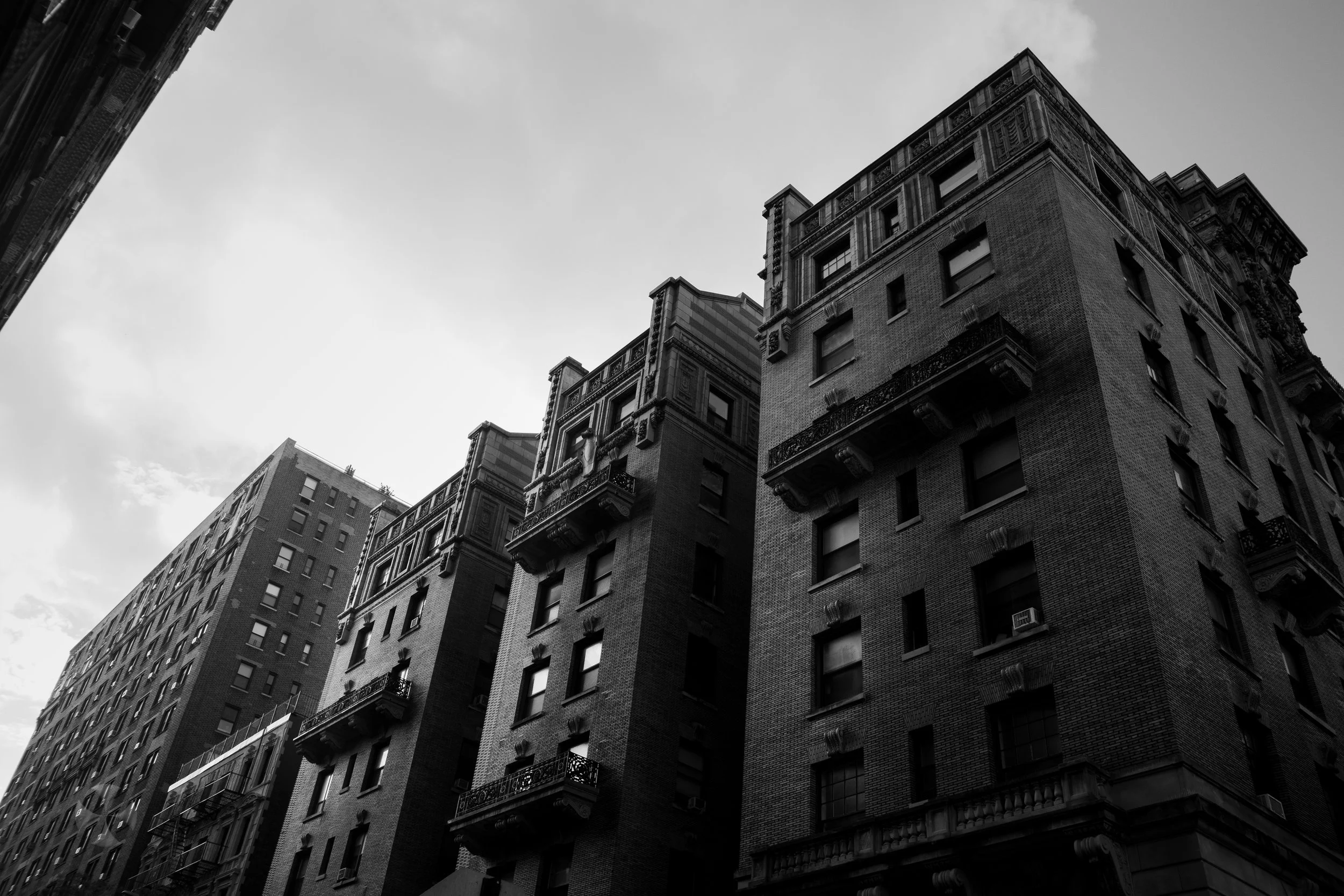 Black and white photo of tall, old buildings with detailed facades and small balconies, viewed from below against a cloudy sky. New York