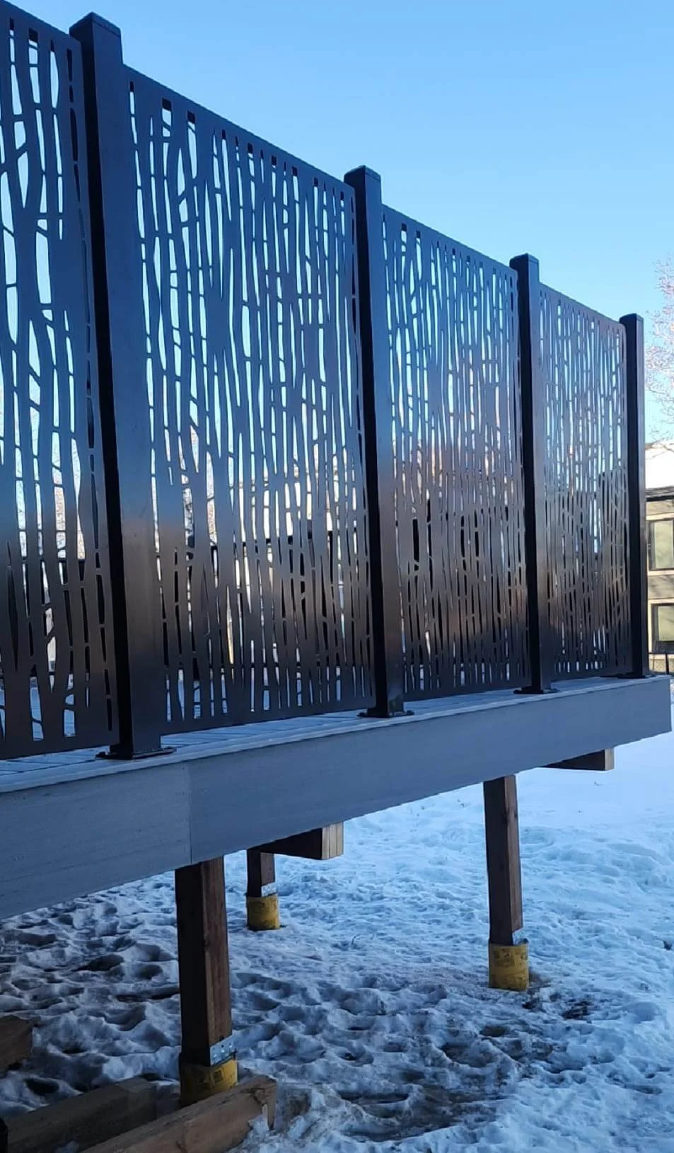 A modern decorative metal railing with tree branch patterns, installed on a raised platform outdoors during winter with snow on the ground.