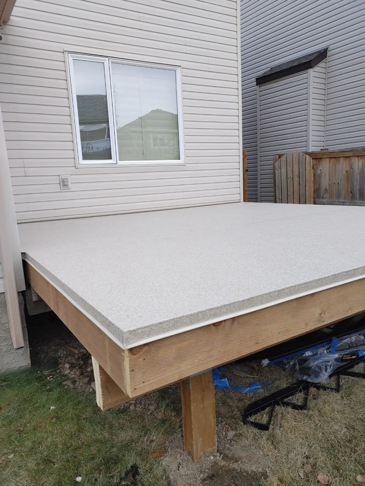 Partially constructed wooden deck with a textured white surface next to a beige house with vinyl siding, adjacent to a gray house with siding, and a small wooden fence in the background.