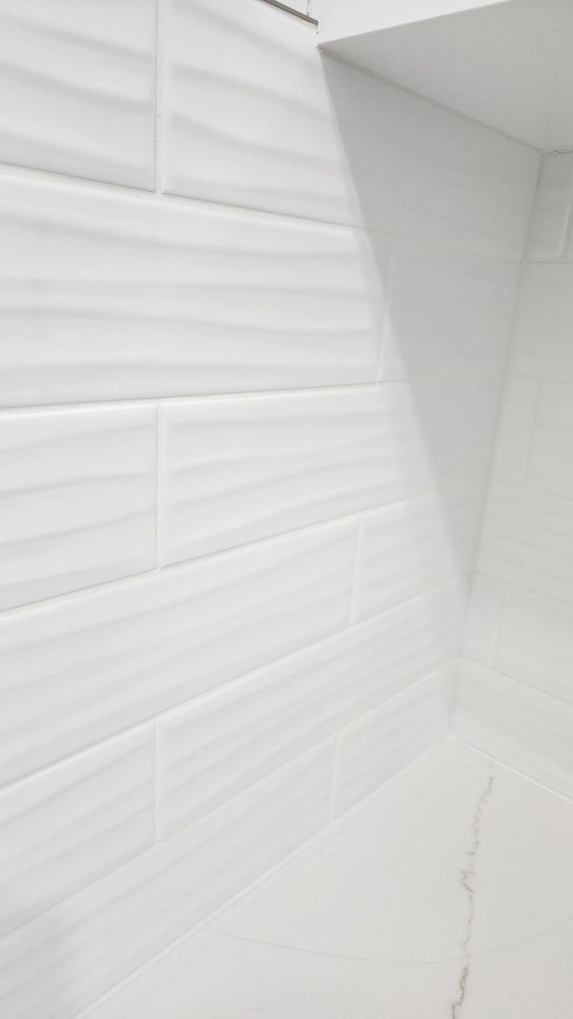 Close-up view of a corner with white tiled walls and a white tiled floor, showing a slight crack and grout lines.