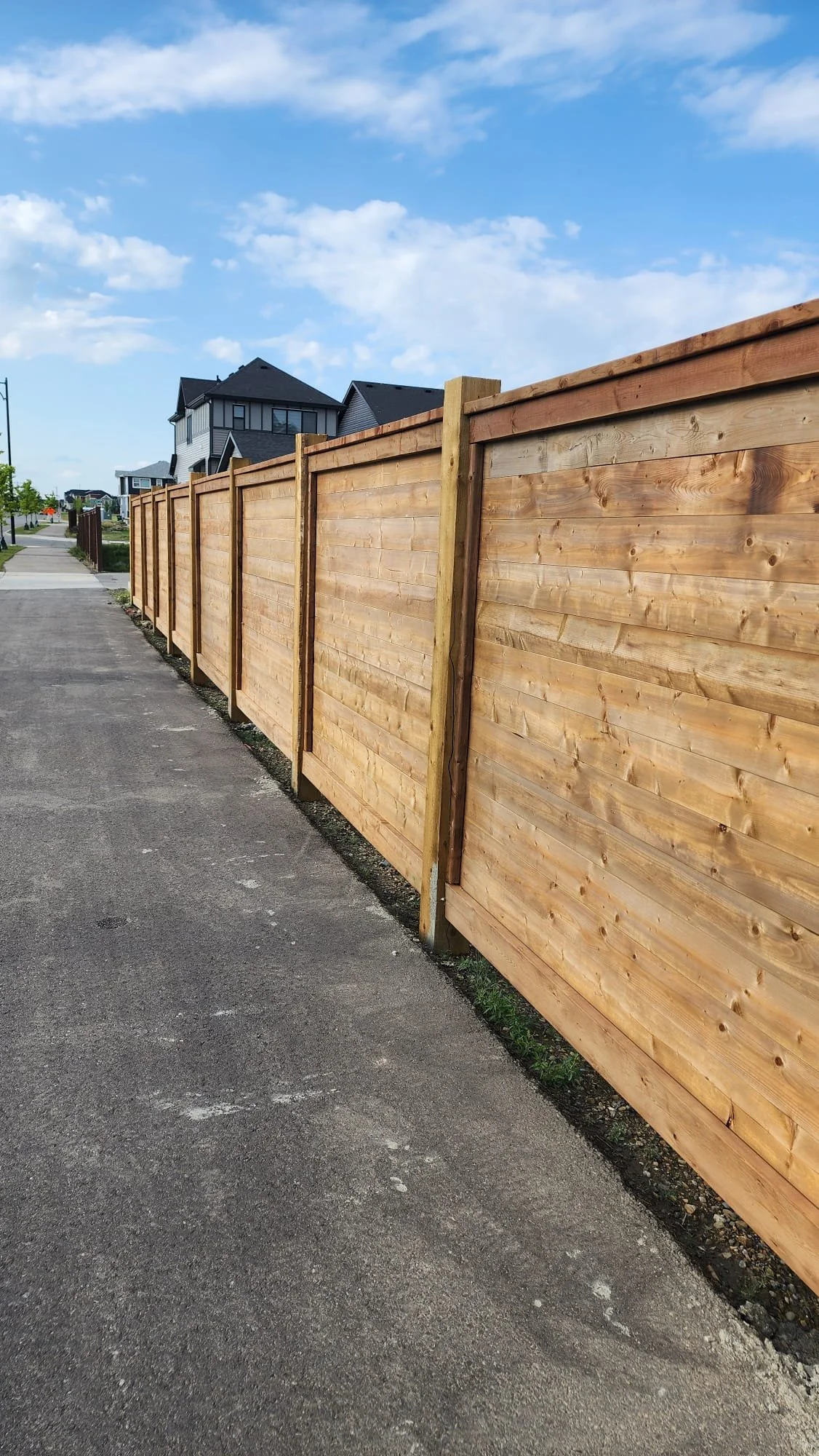 A freshly built wooden fence runs along the sidewalk in a suburban neighborhood under a partly cloudy sky.