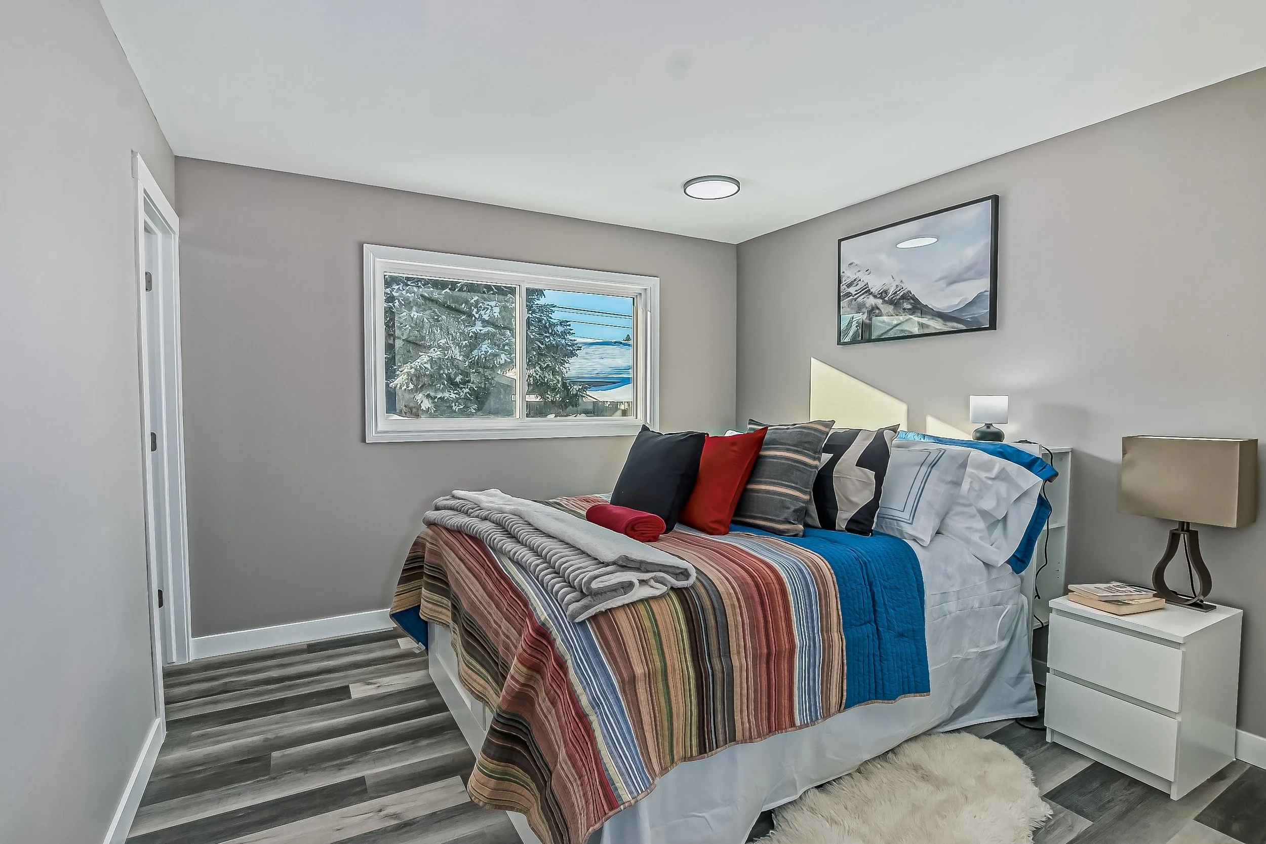 Bedroom with a large bed, striped quilt, numerous pillows, side tables with lamps, a framed mountain landscape picture, and a window with a snowy outdoor view.