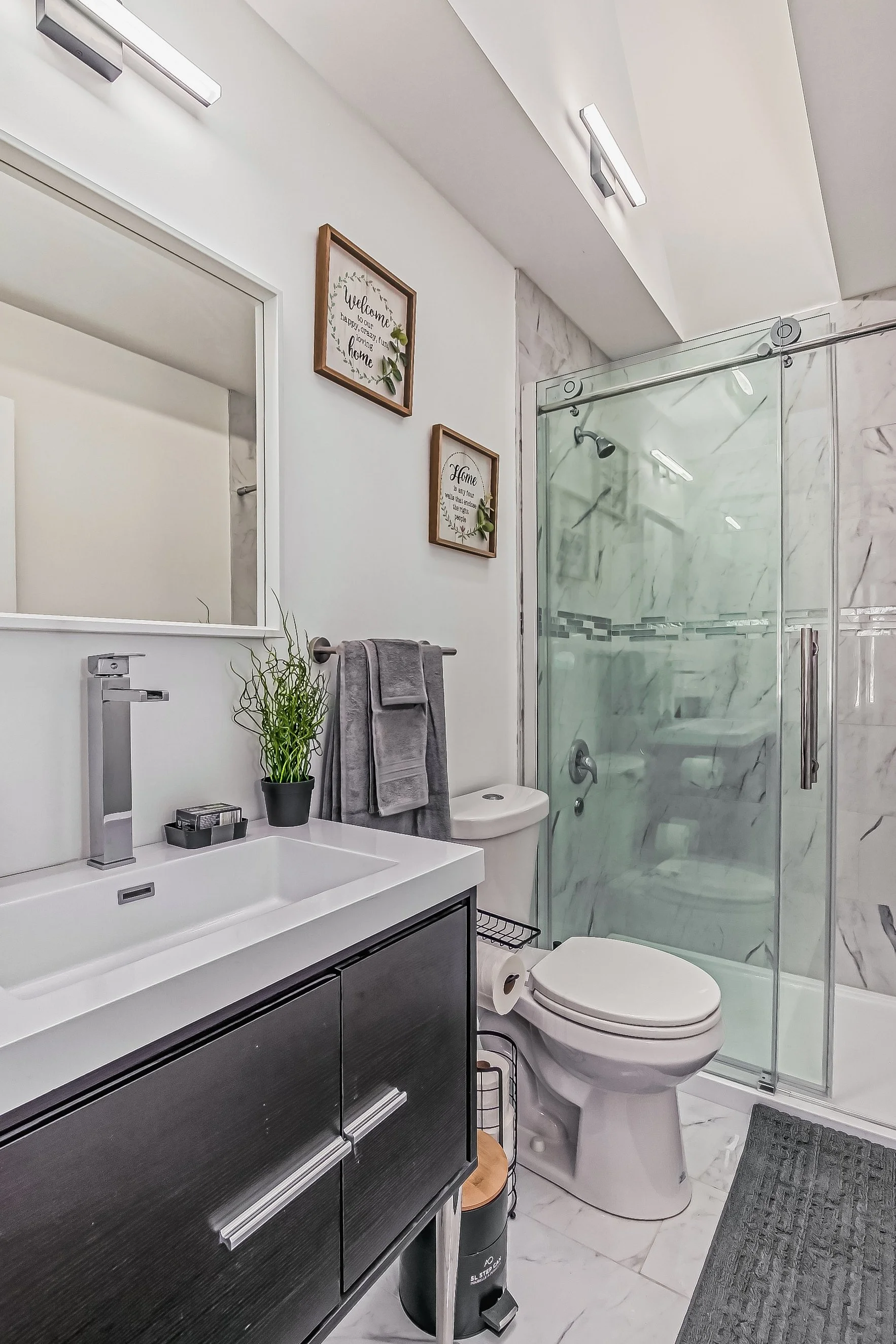 Modern bathroom with white walls, a large mirror, a black vanity with a white sink, a potted plant, gray towels, a toilet paper holder, framed wall art, and a glass shower enclosure with marble tiles.