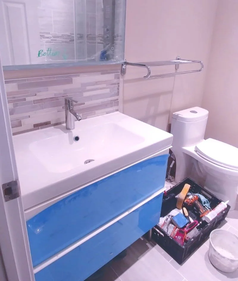 Bathroom with a white sink, a blue cabinet beneath, a mirror above, a toilet to the right, and a black toolbox filled with tools on the floor. A towel rack is mounted on the wall above the sink.