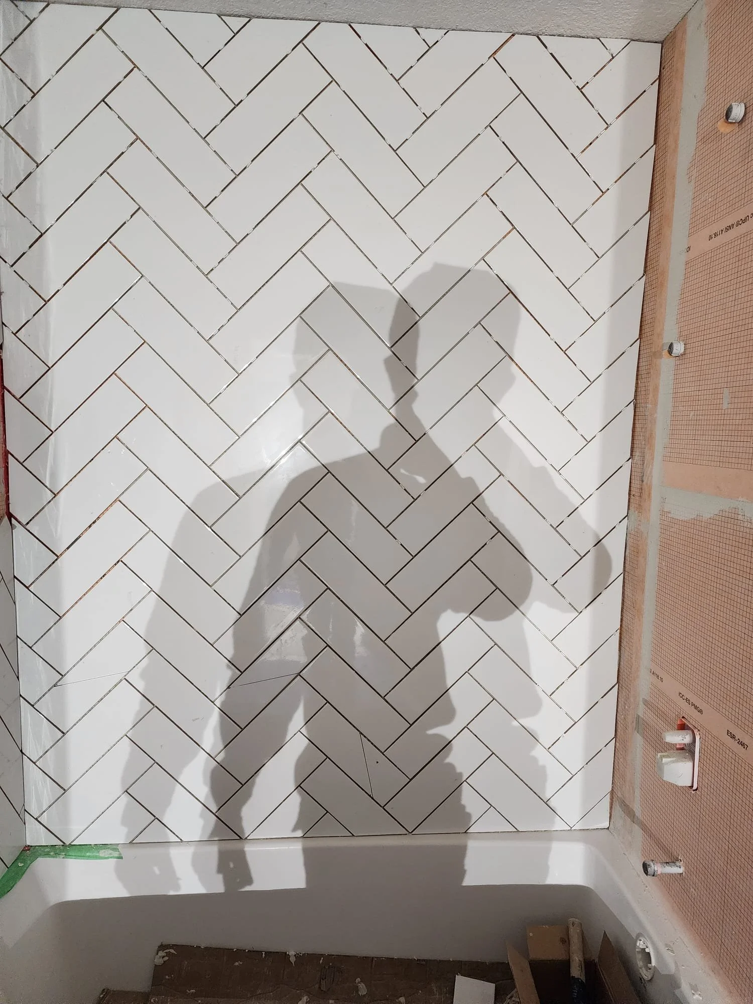 Shadow of a person taking a photo is cast on a white herringbone tile bathroom wall, with unfinished areas and plumbing fixtures visible.
