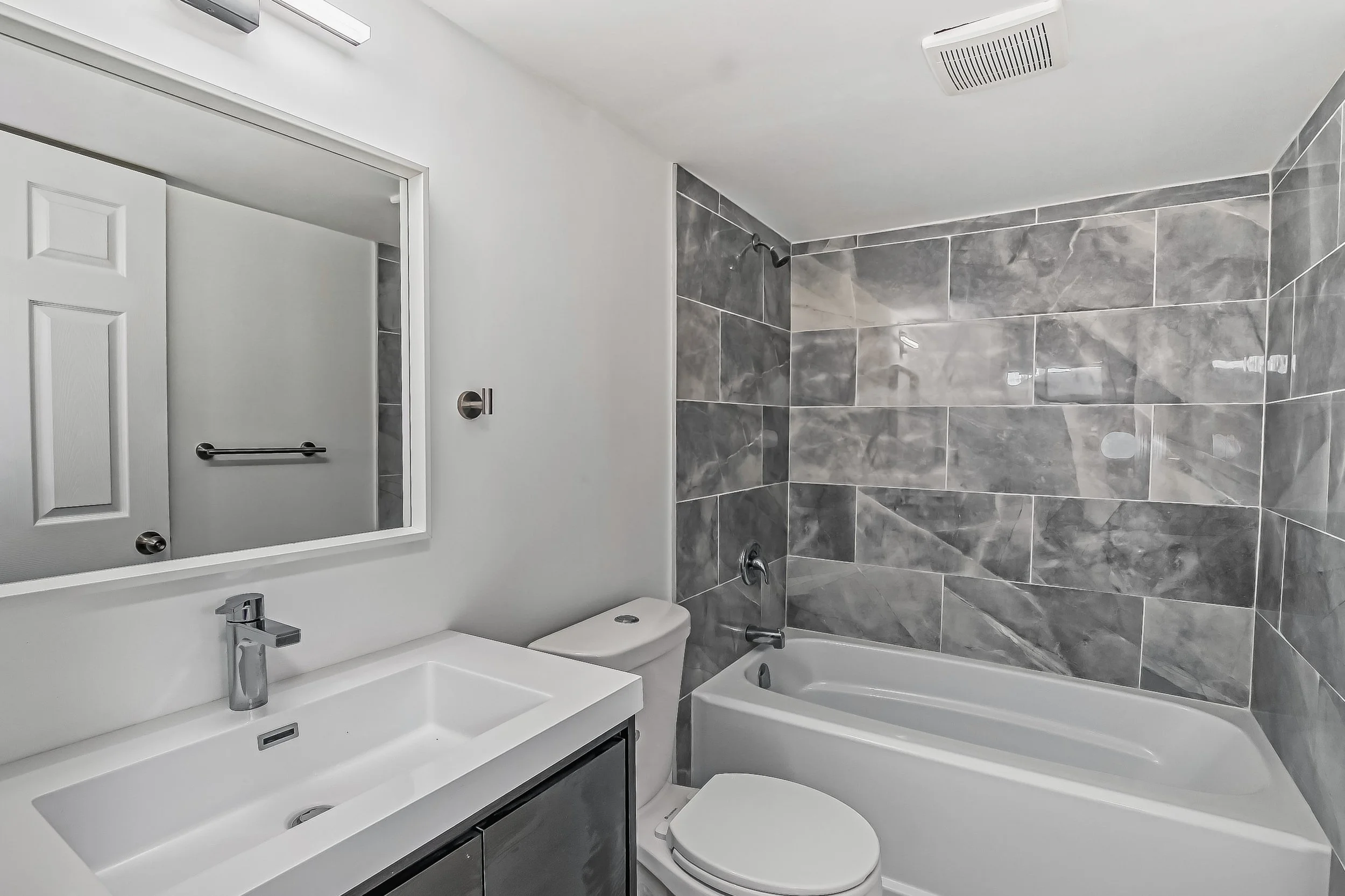 Modern bathroom with white sink, mirror, toilet, and bathtub with gray marble tile walls.