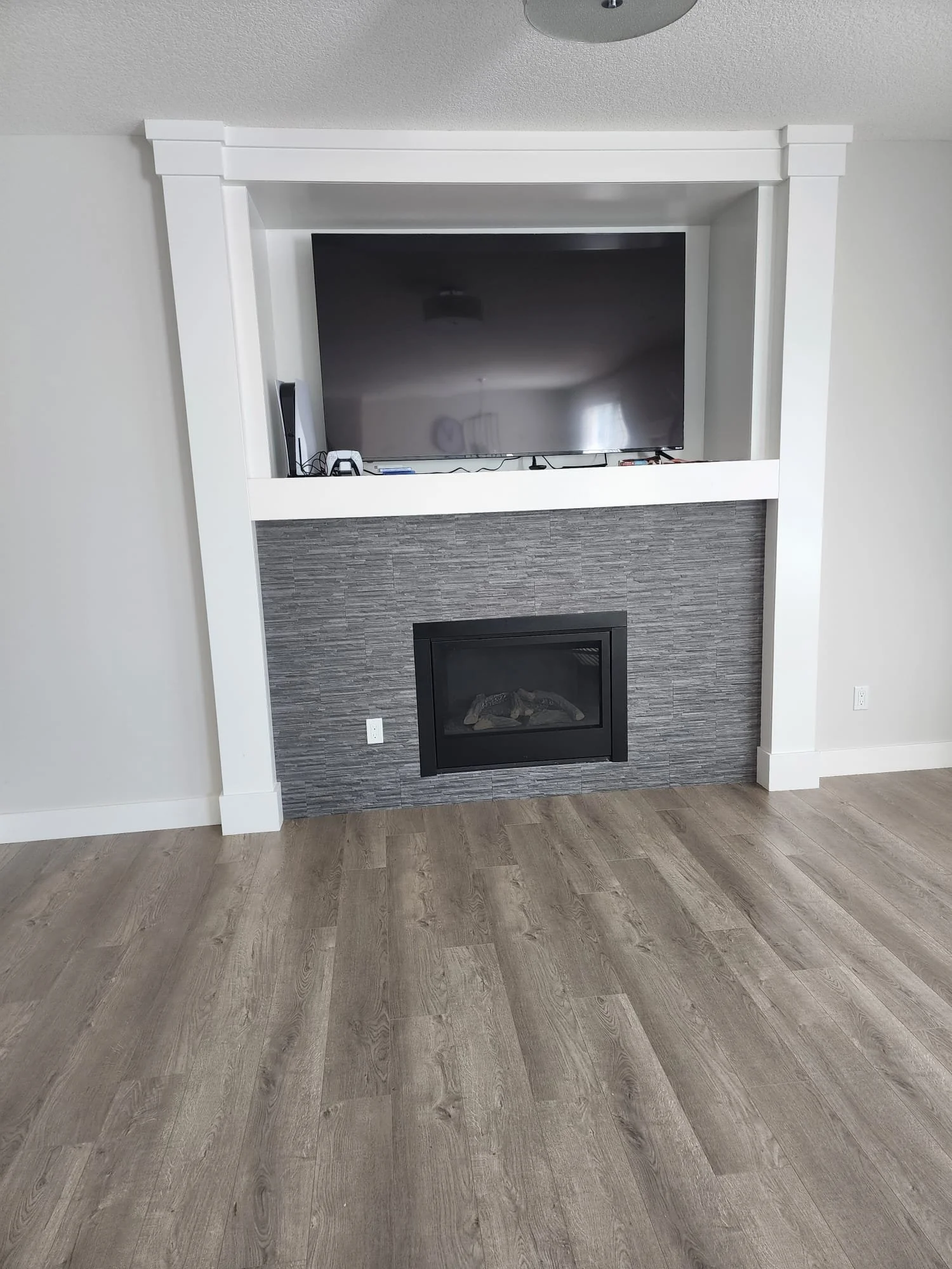 Living room with a fireplace and a mounted flat-screen TV above it. The wall around the fireplace is decorated with gray textured tiles, and the room has light-colored walls and wood flooring.