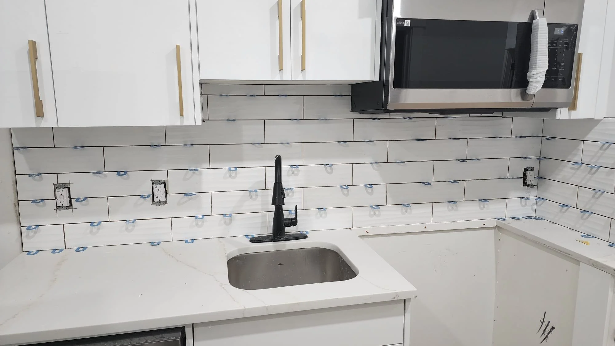 Kitchen with white cabinets, black faucet, white marble countertop, tile backsplash, and a microwave. Some electrical outlets are visible without cover plates.