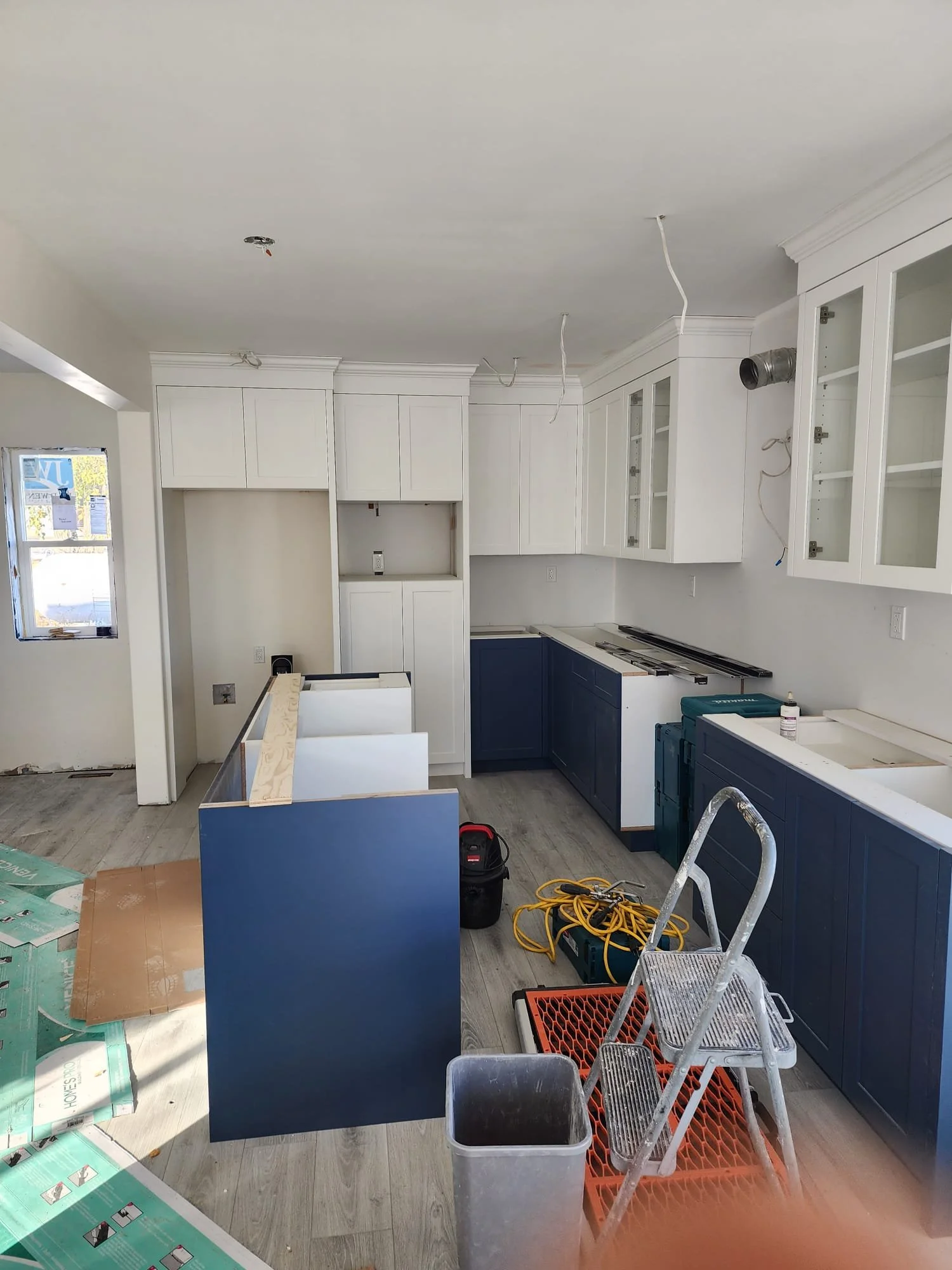 Kitchen under renovation with white upper cabinets, partially installed dark blue lower cabinets, construction tools, and a step ladder.