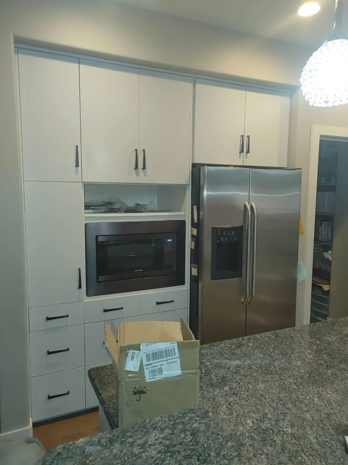 Kitchen with white cabinets, stainless steel microwave, and double-door refrigerator. There is a cardboard box on a granite countertop.