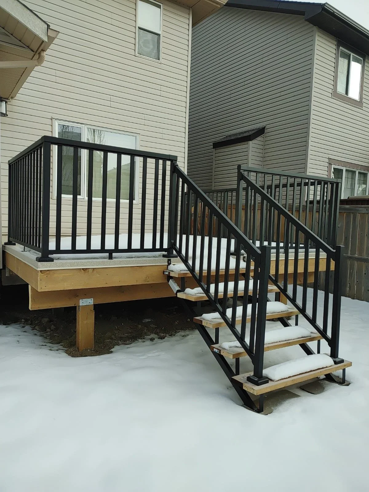 Newly built wooden deck with black metal railing and staircase in a snowy backyard, attached to a beige house with vinyl siding.