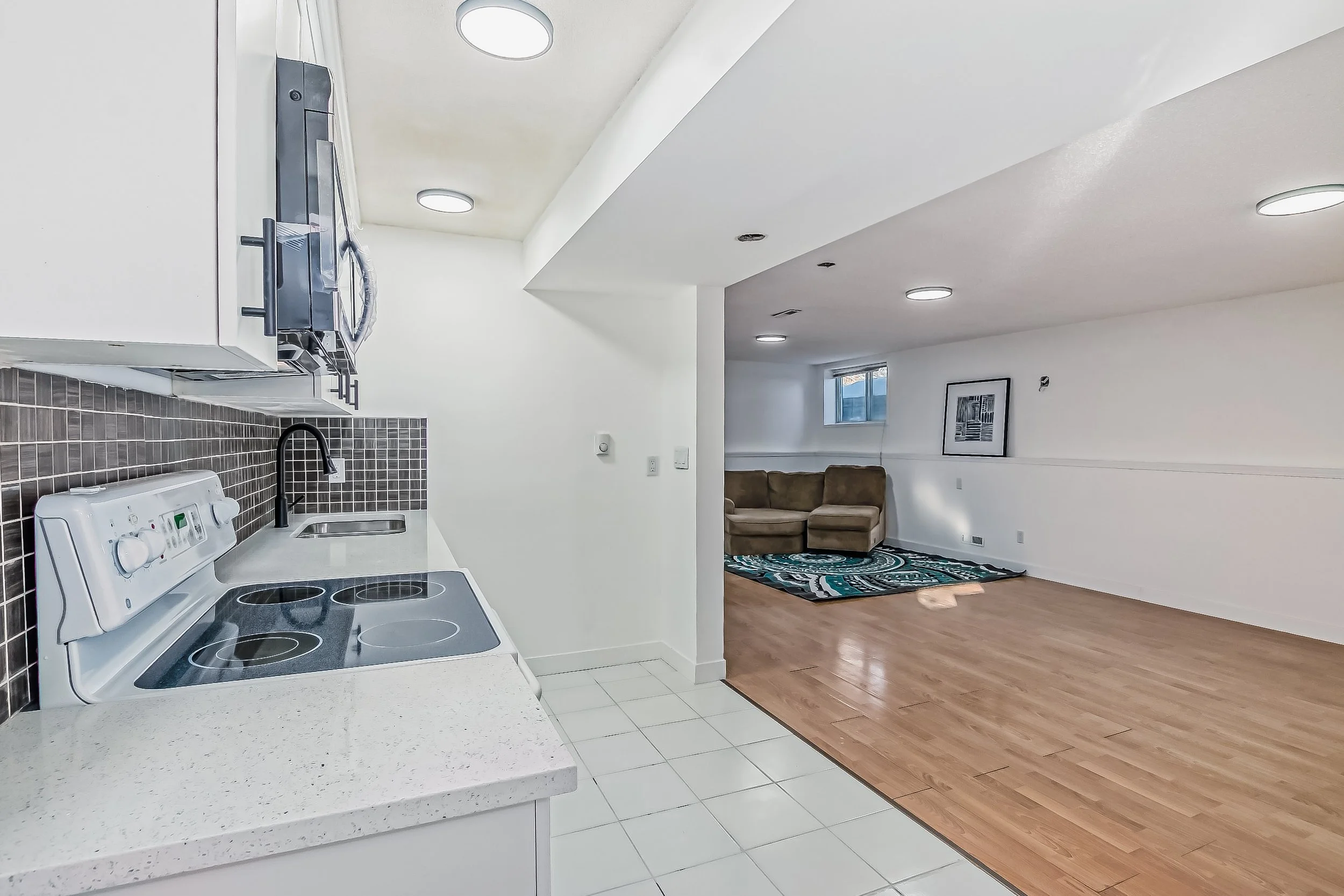 View of a basement living room with a small kitchen area on the left, featuring white cabinets, a black and white speckled counter, a black sink, a black microwave, and a white stove. The living area has a brown sectional sofa, a colorful abstract ru