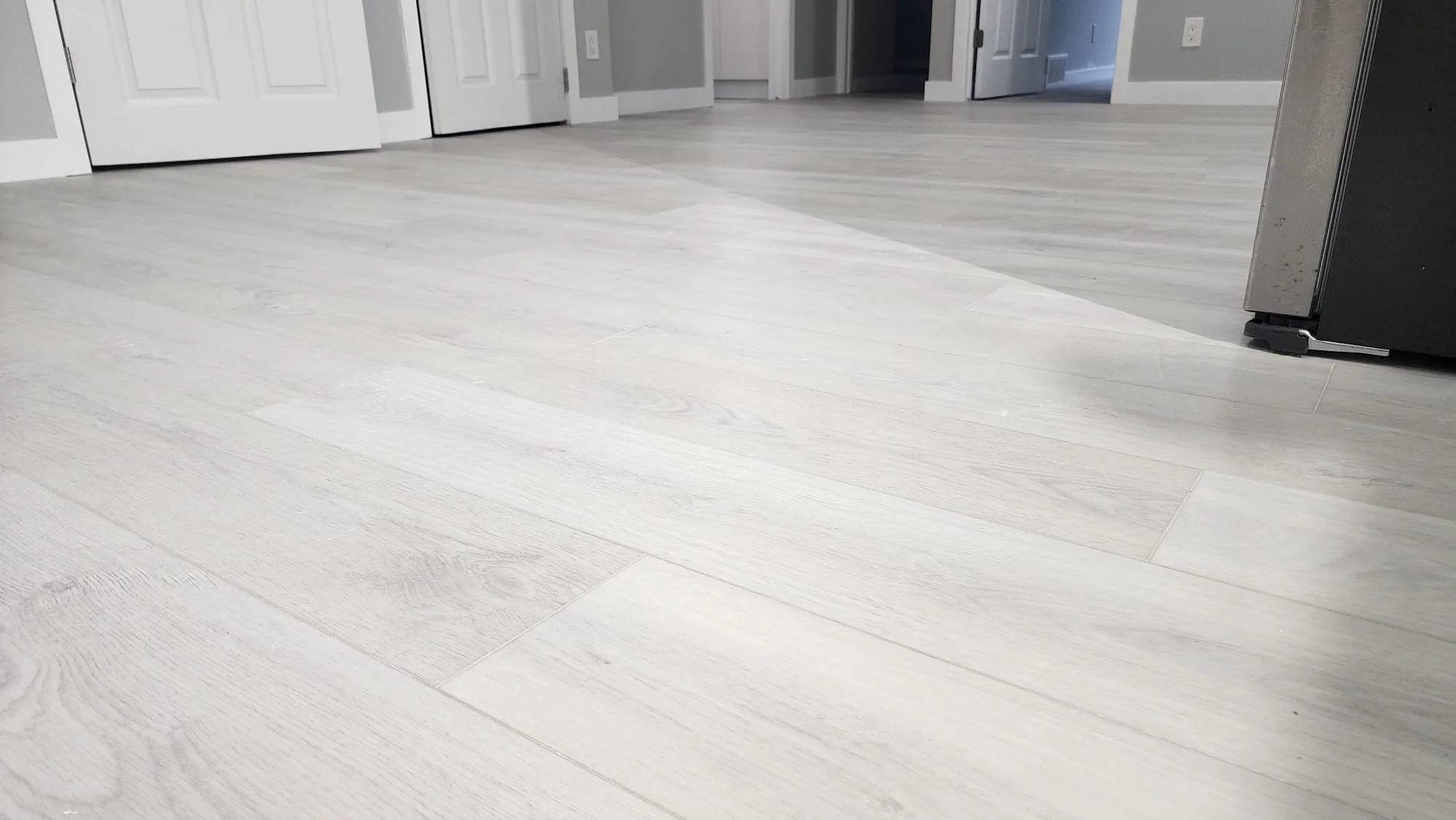 Light-colored wooden flooring in a room with visible doors, appliances, and walls.