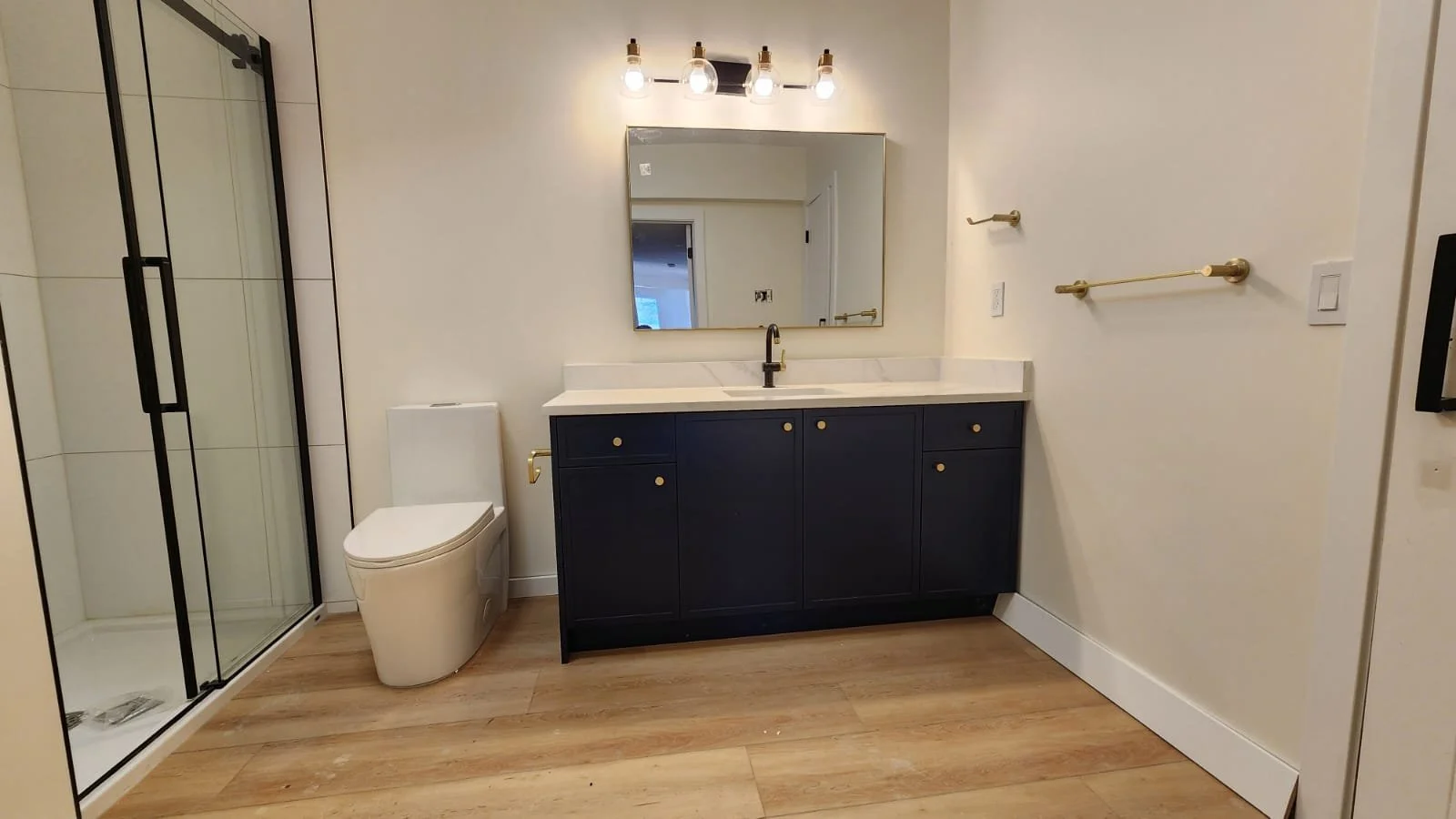 Modern bathroom with a walk-in shower, white toilet, navy blue vanity with gold hardware, marble countertop, and a large mirror, with wood flooring and gold towel bars.
