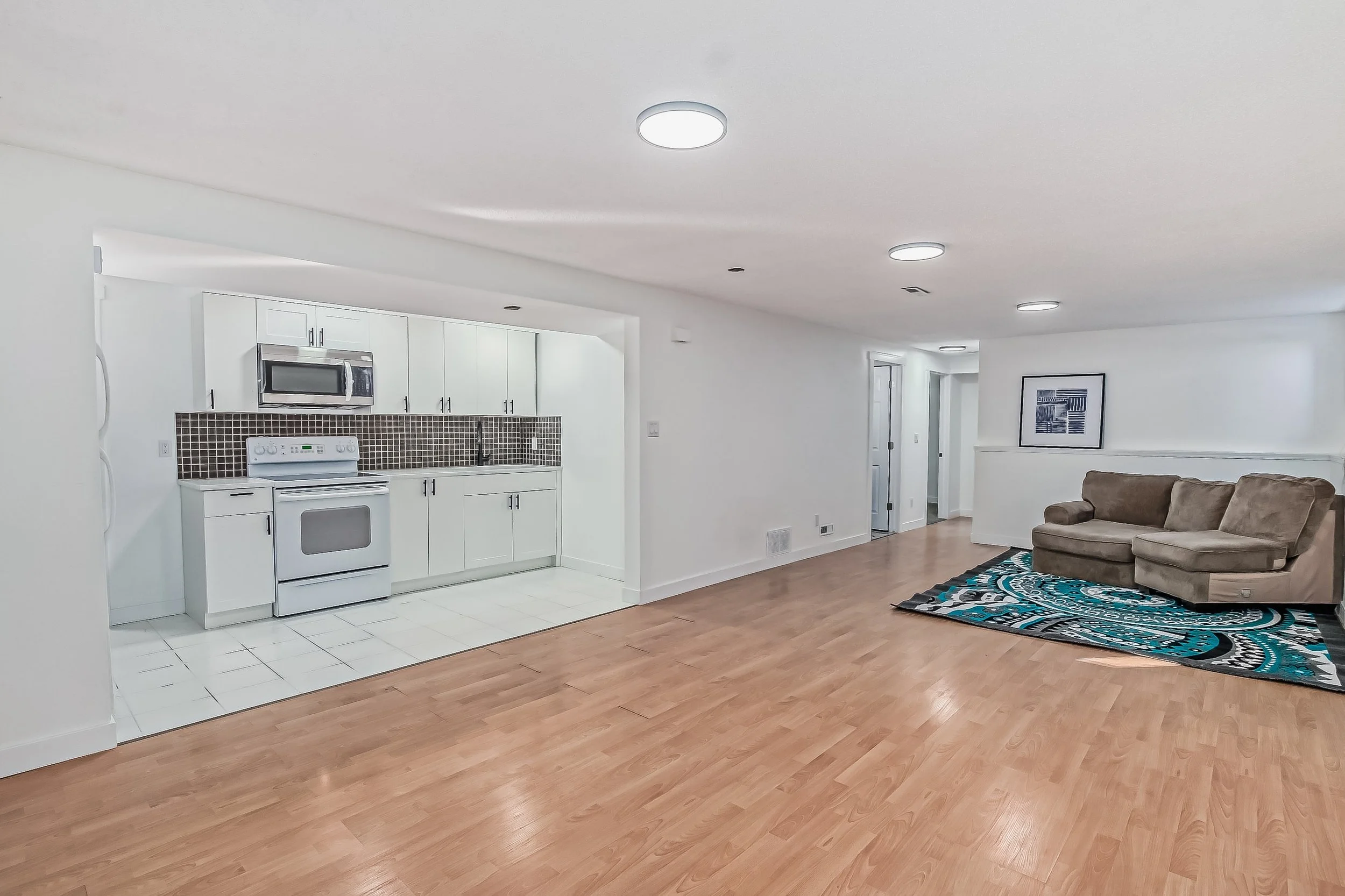 Open-concept living area with a small kitchen featuring white cabinets, a brown tiled backsplash, and white appliances. A beige sofa is positioned on a colorful, patterned rug, with light hardwood flooring throughout the space.