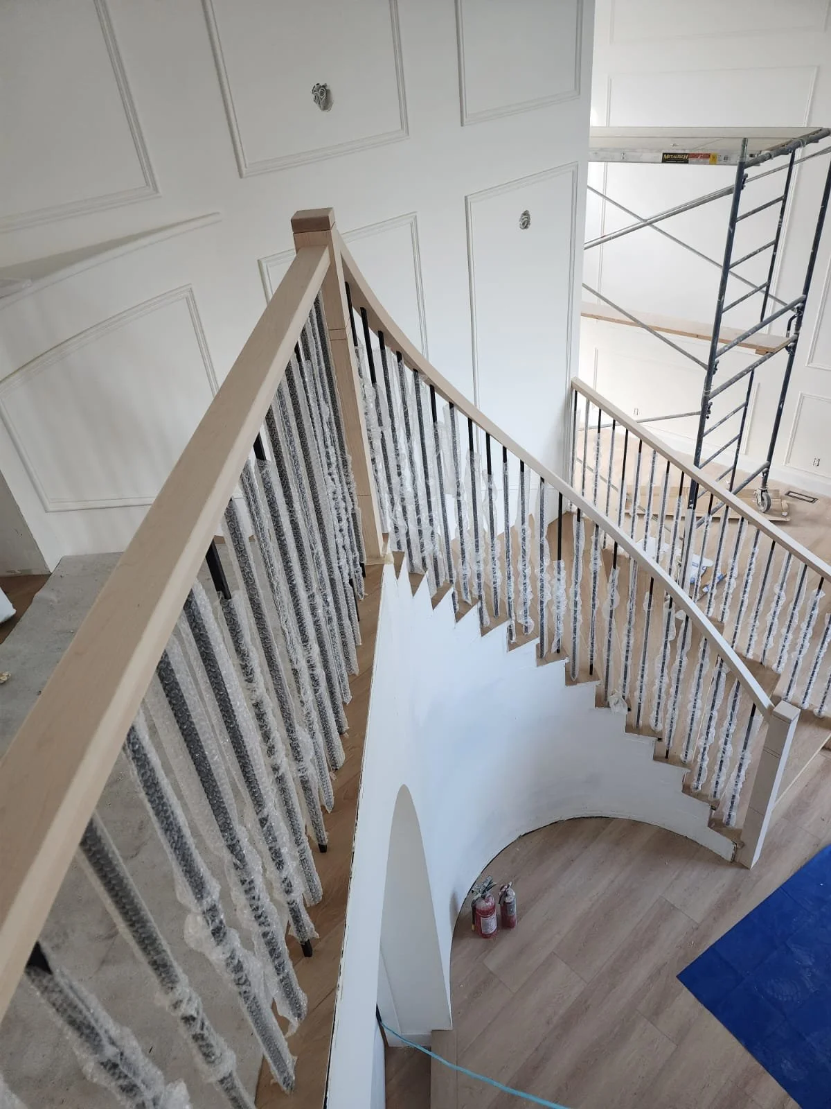 Interior view of a staircase under construction with a wooden handrail, partially covered winders, and scaffolding in the background.