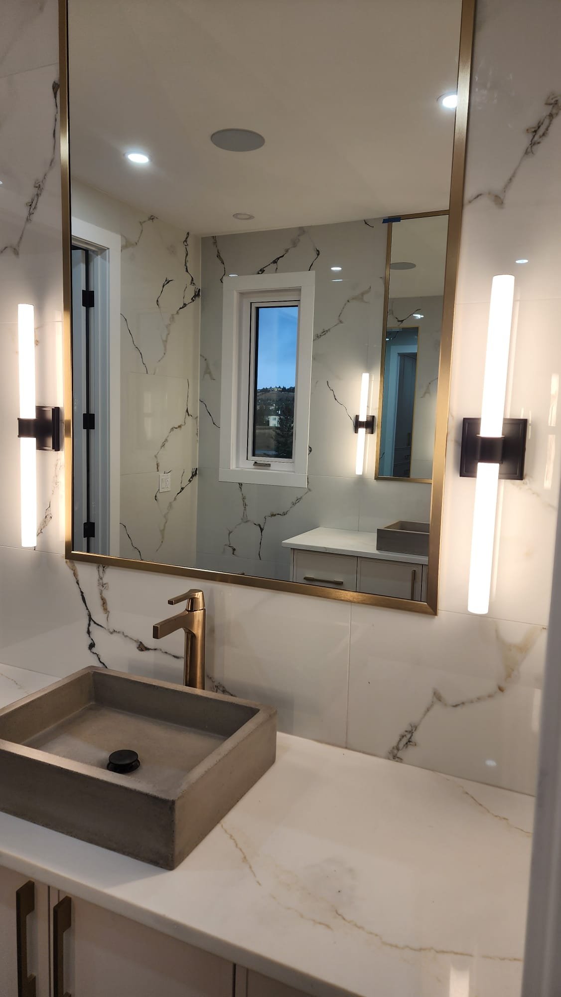 Modern bathroom with white marble walls and countertop, rectangular concrete sink, gold faucet, large mirror with gold trim, and vertical light fixtures on both sides of the mirror. There is a small window showing an outdoor view.