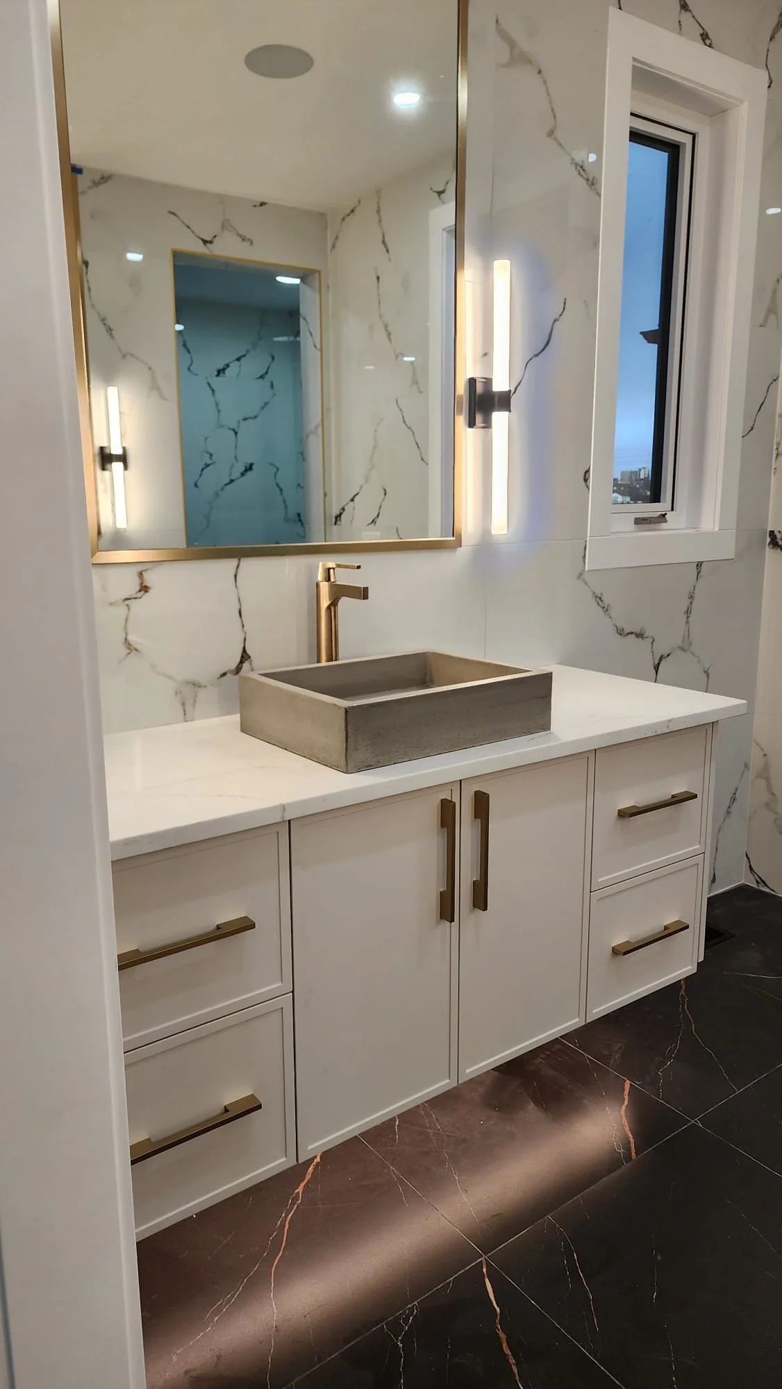 Modern bathroom vanity with white cabinets, gold handles, a concrete sink, gold faucet, large mirror, and marble wall tiles. There is a window and wall-mounted lights.