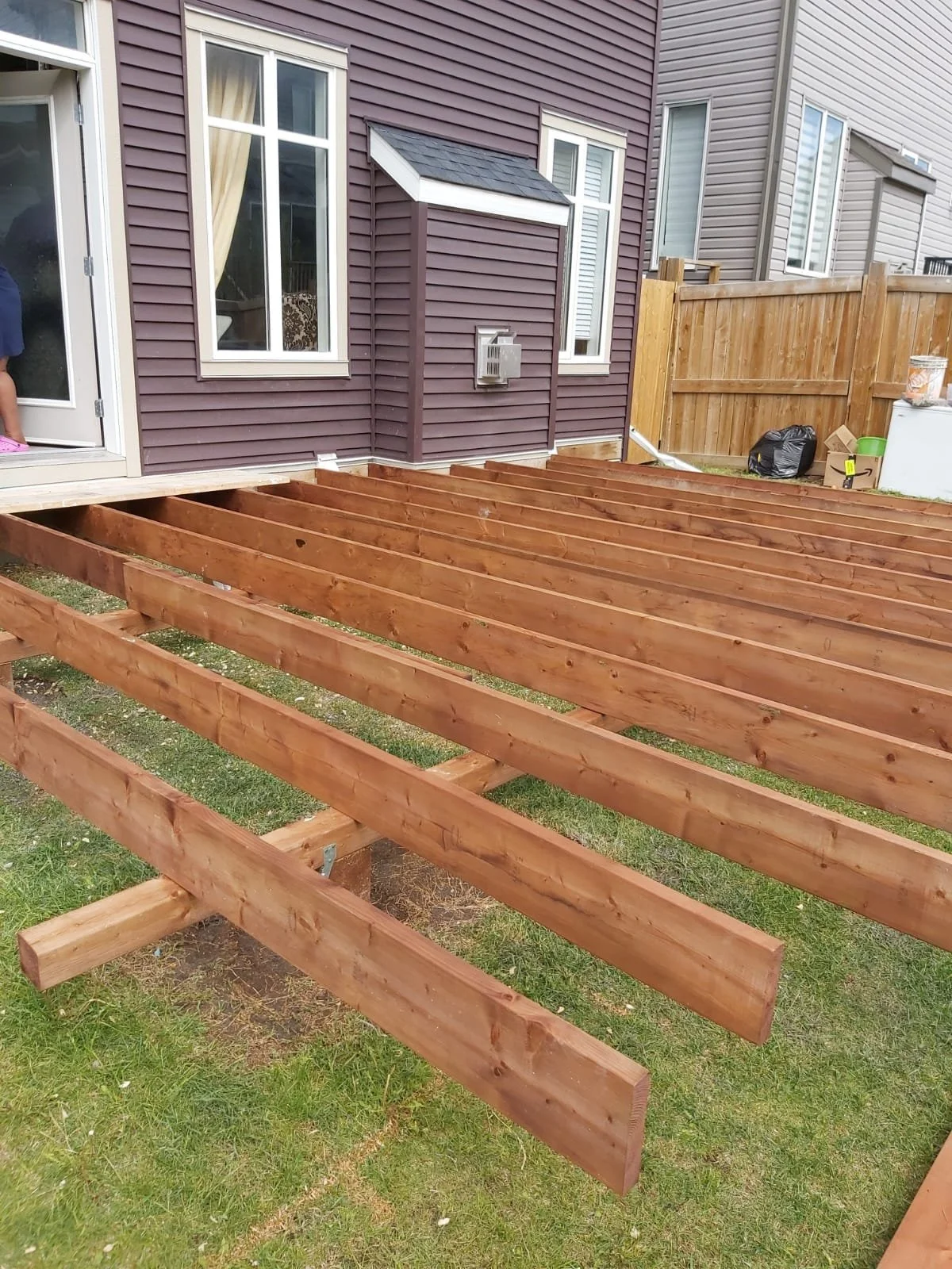 Construction of a wooden deck outside a house with purple and gray siding, with empty wooden beams laid out on the grass.