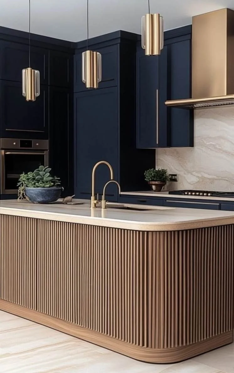 Modern kitchen with navy blue cabinets, gold pendant lights, a beige curved island with wooden slats, a built-in oven, green plants, and a marble backsplash.