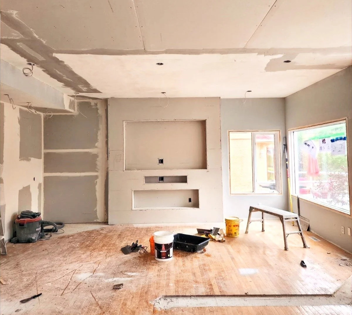Interior room under renovation with drywall installation, construction tools, and unfinished surfaces, including wires hanging from the ceiling and windows providing natural light.