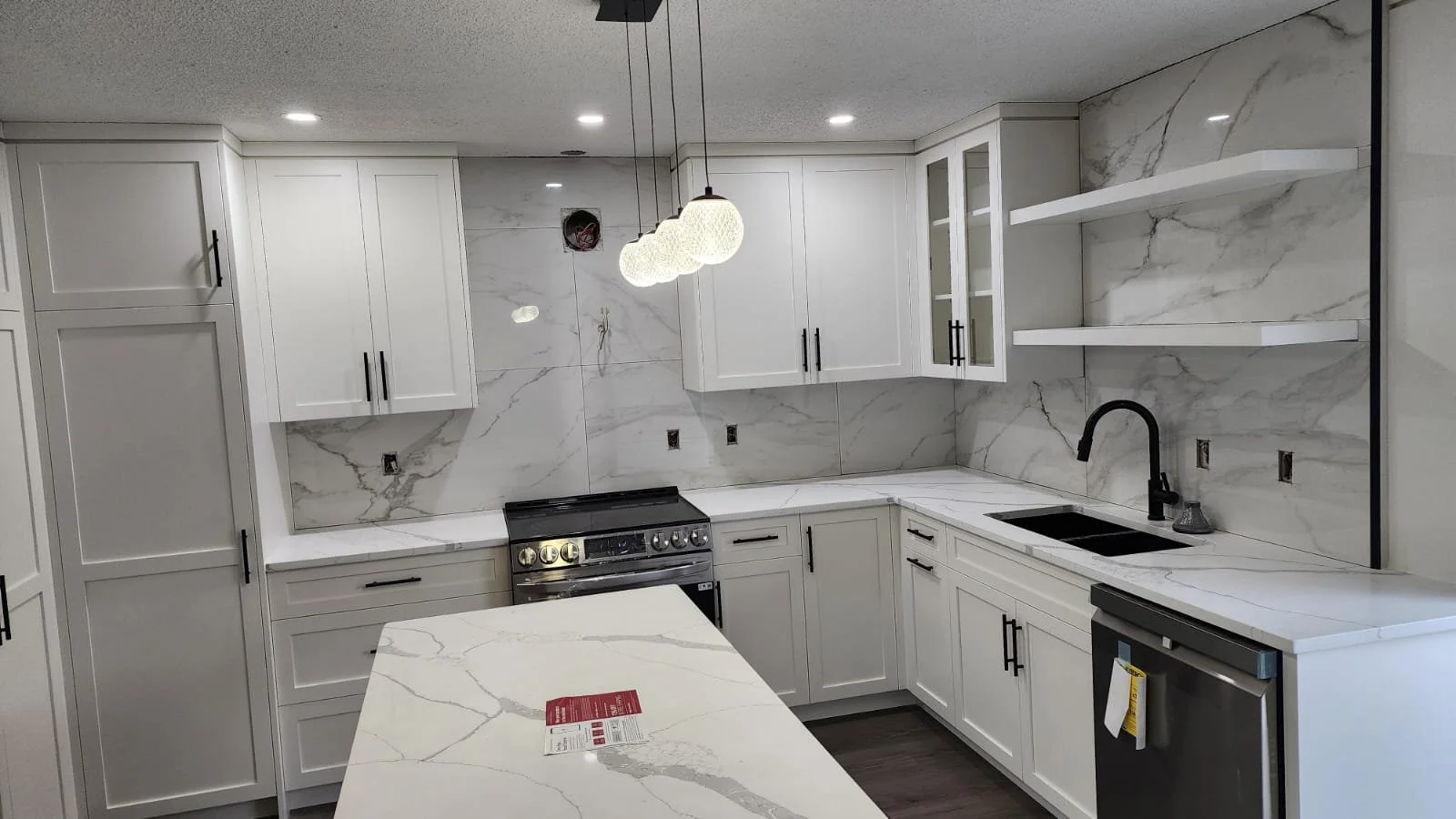 Modern kitchen with white cabinets, marble countertops, black hardware, a black sink faucet, and stainless steel appliances, including an oven and dishwasher. There are pendant lights hanging above the island.