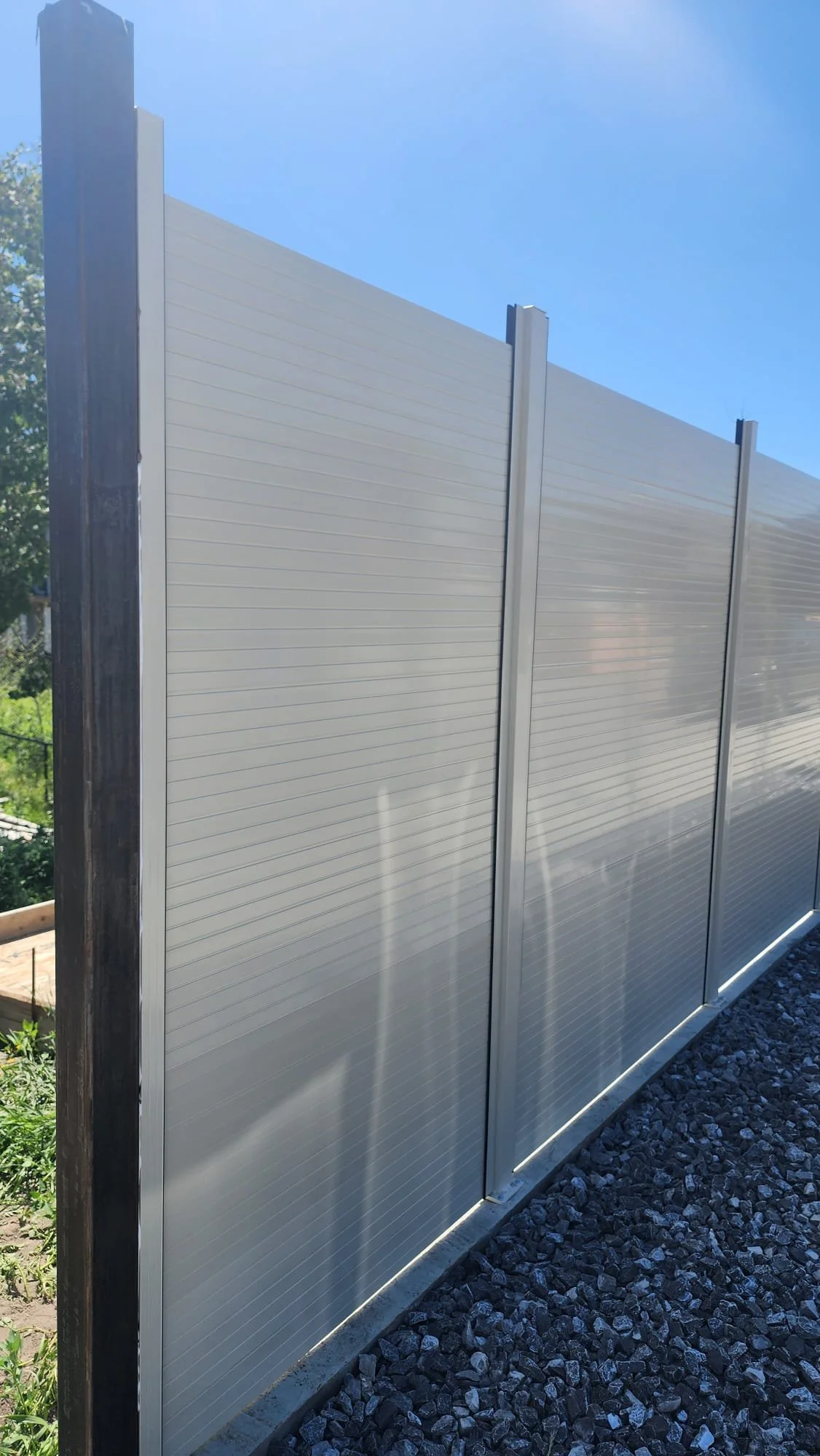 White metal fence panels supported by vertical posts, beside a gravel ground, under a blue sky.