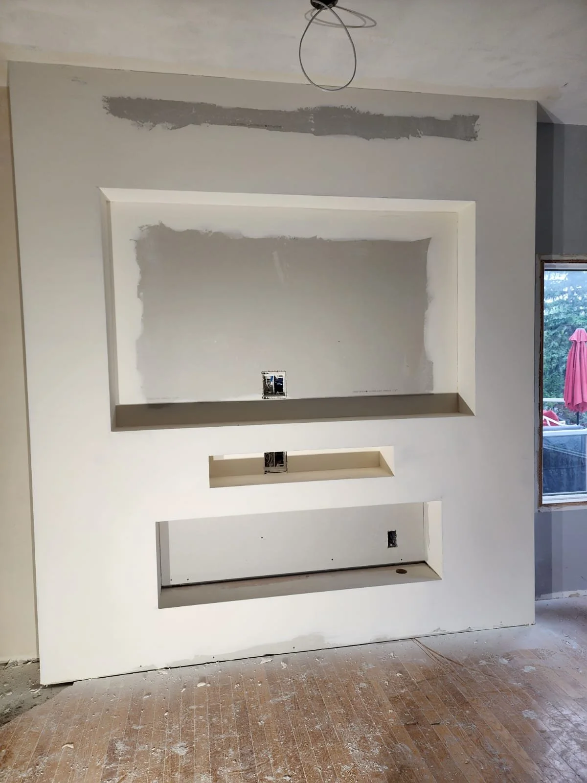 Interior of a room under construction with a large wall featuring three recessed wall niches, electrical outlets, and unfinished drywall with visible joints and mud, and a window to the right showing outdoor trees and a patio umbrella.