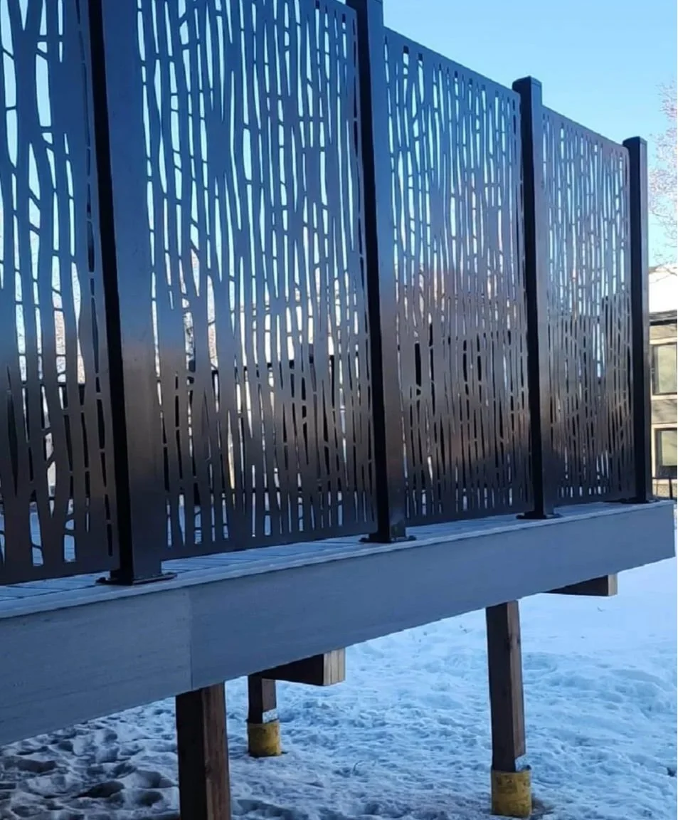 Close-up of a decorative metal railing with a tree branch pattern, set on a wooden deck in a snowy outdoor setting.