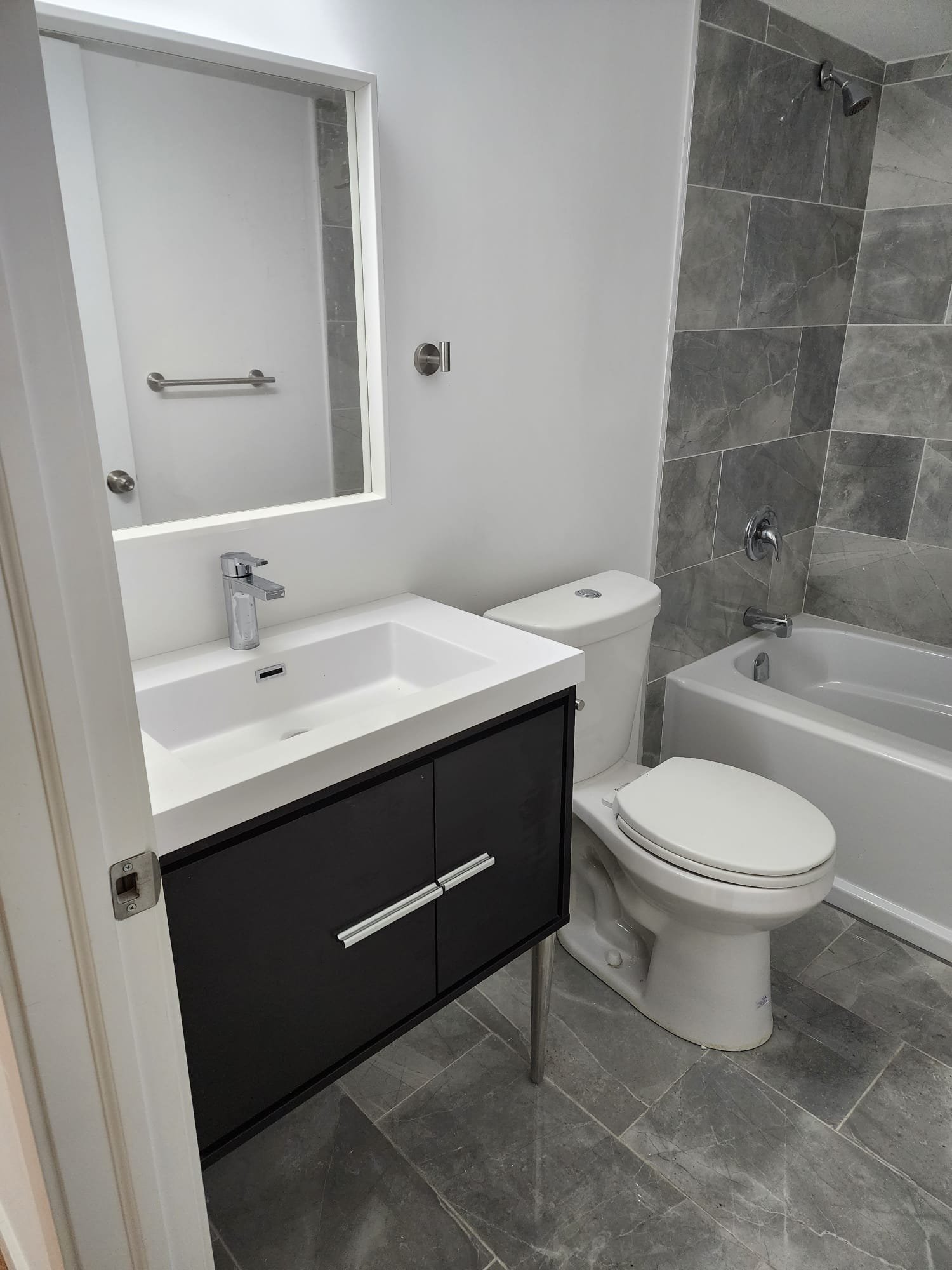 Modern bathroom featuring a white vanity with a black cabinet, a mirror above the sink, a toilet, and a bathtub with a gray tiled wall.